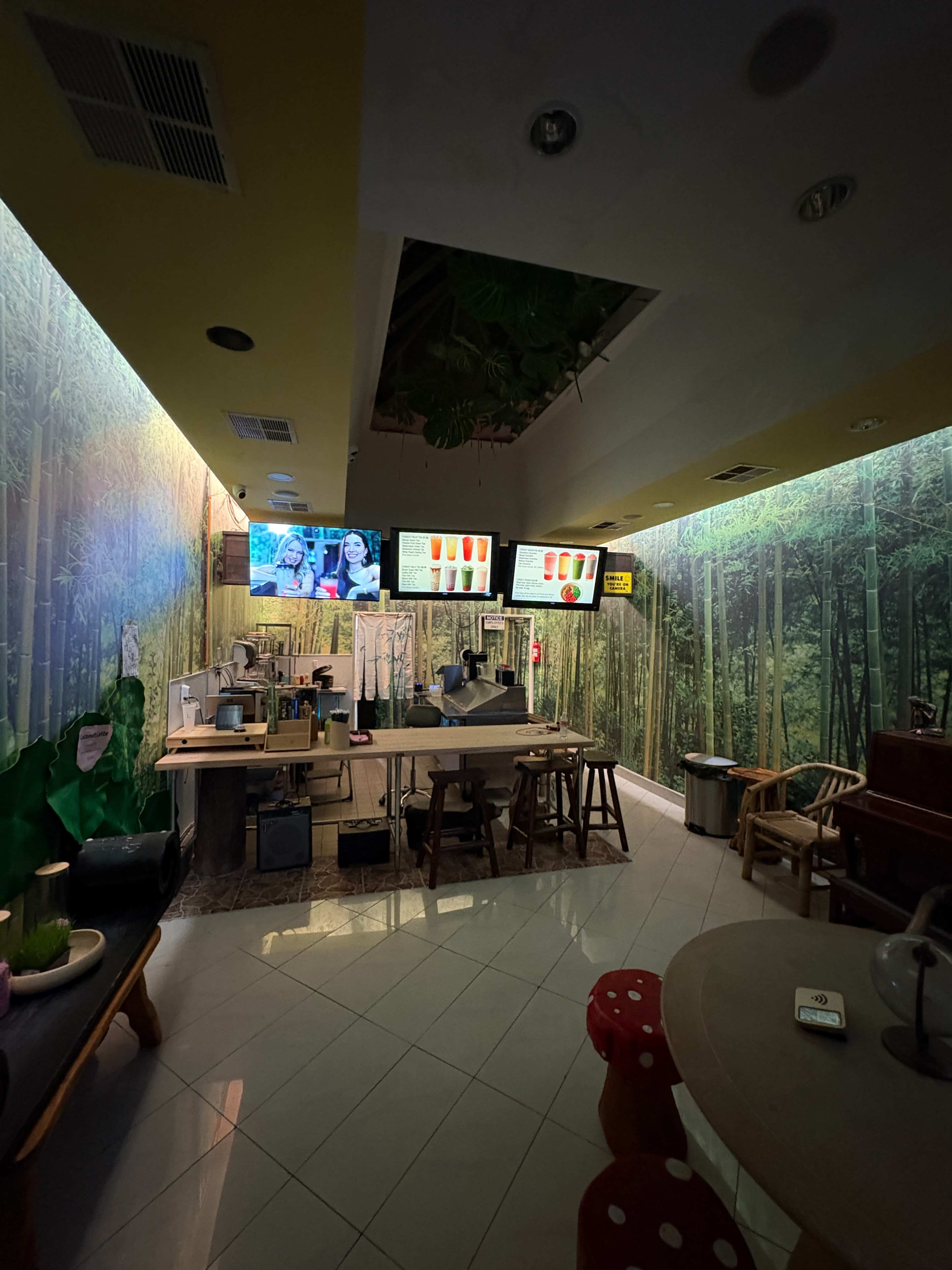 The image shows a spacious café interior with a bamboo-themed mural, several screens displaying menu items, and a counter area equipped with seating.