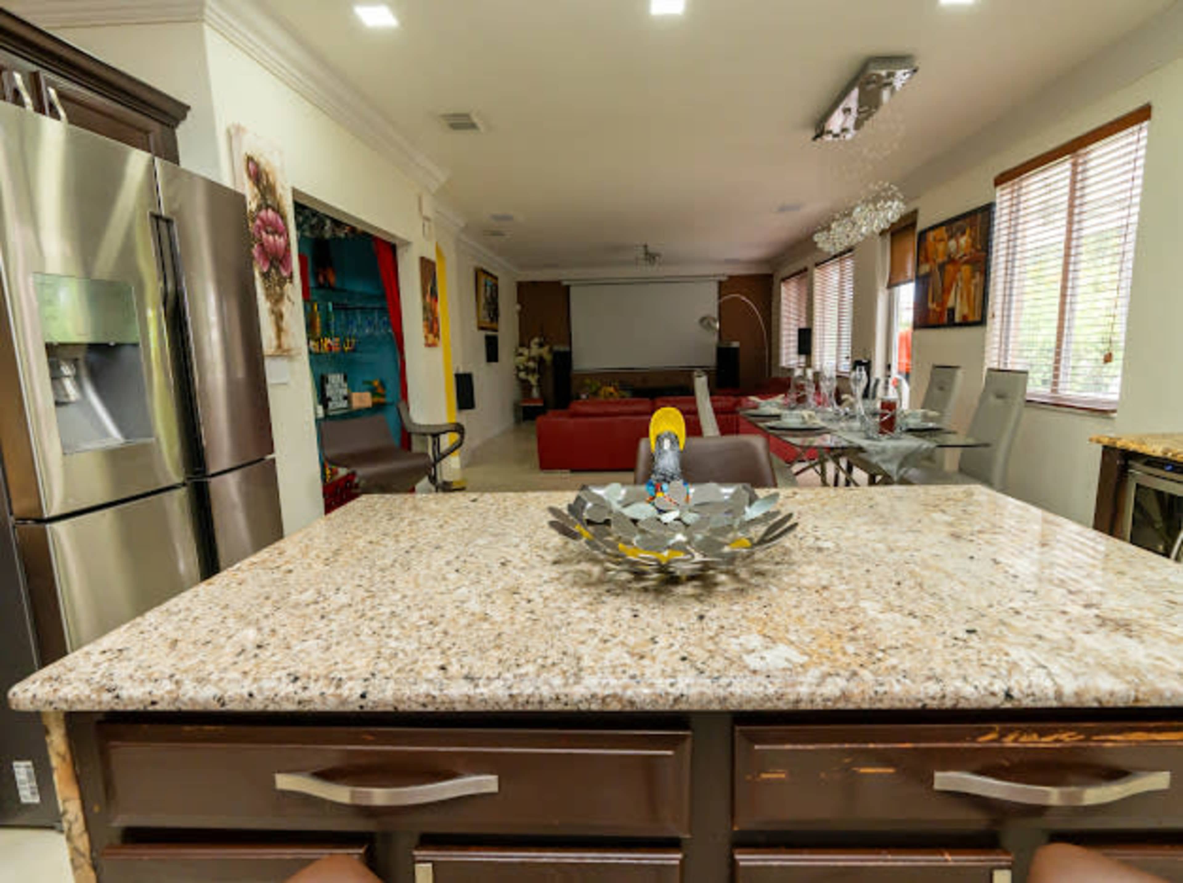 A modern kitchen featuring a granite countertop with a decorative bowl, overlooking a spacious living area with a large red couch and a home theater setup.