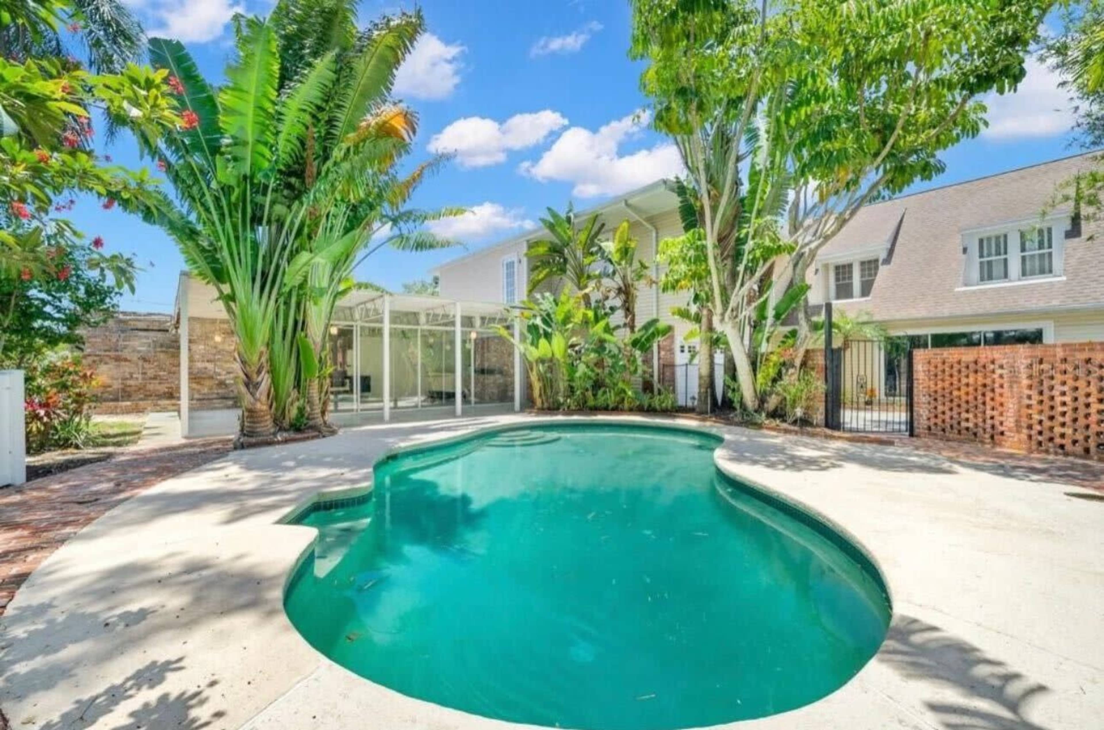 The image shows a swimming pool surrounded by tropical plants and a landscaped outdoor area next to a house.