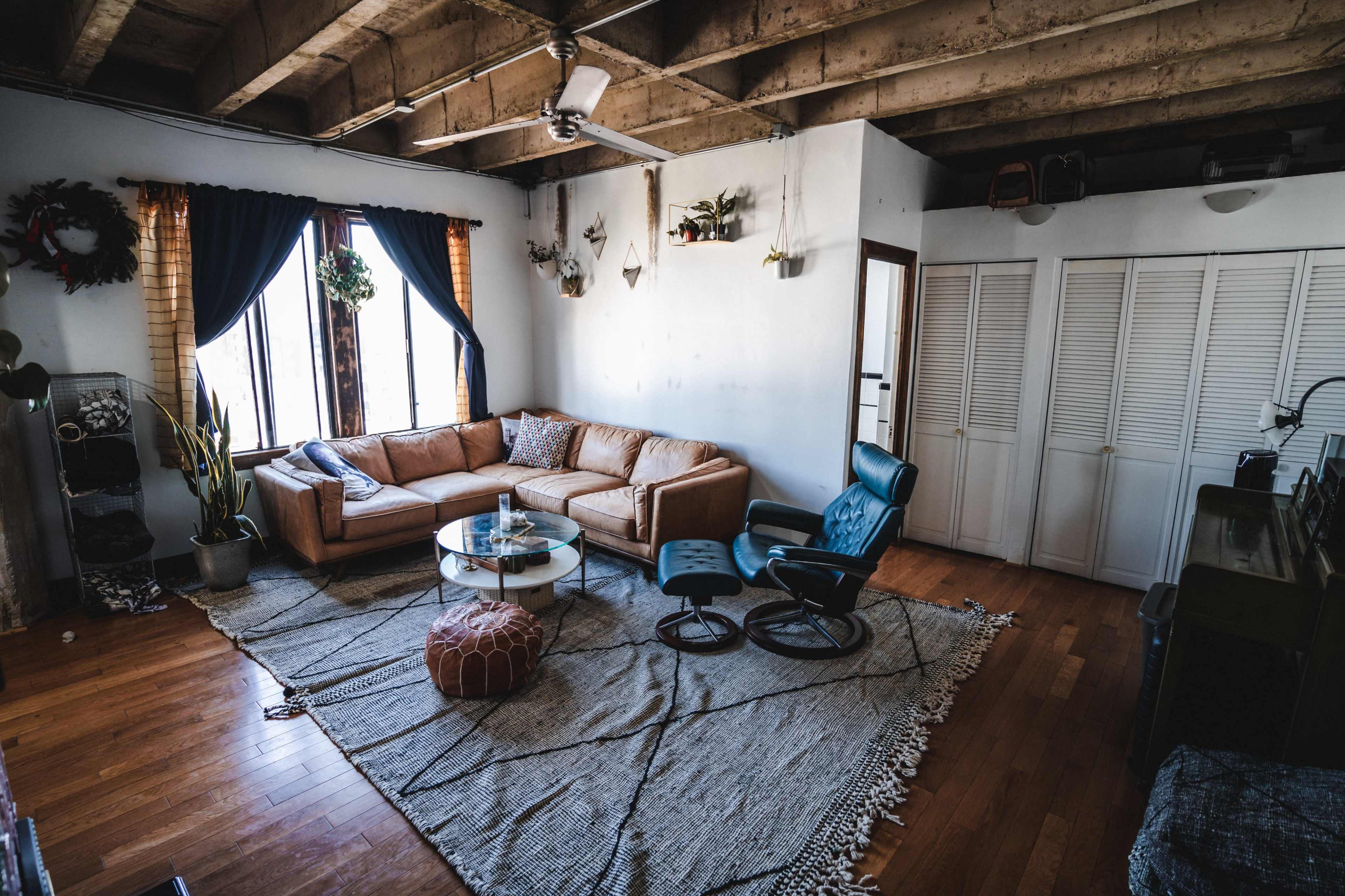 The image shows a cozy living room with a brown sectional couch, a round glass coffee table, a blue lounge chair, and a patterned area rug on hardwood flooring.