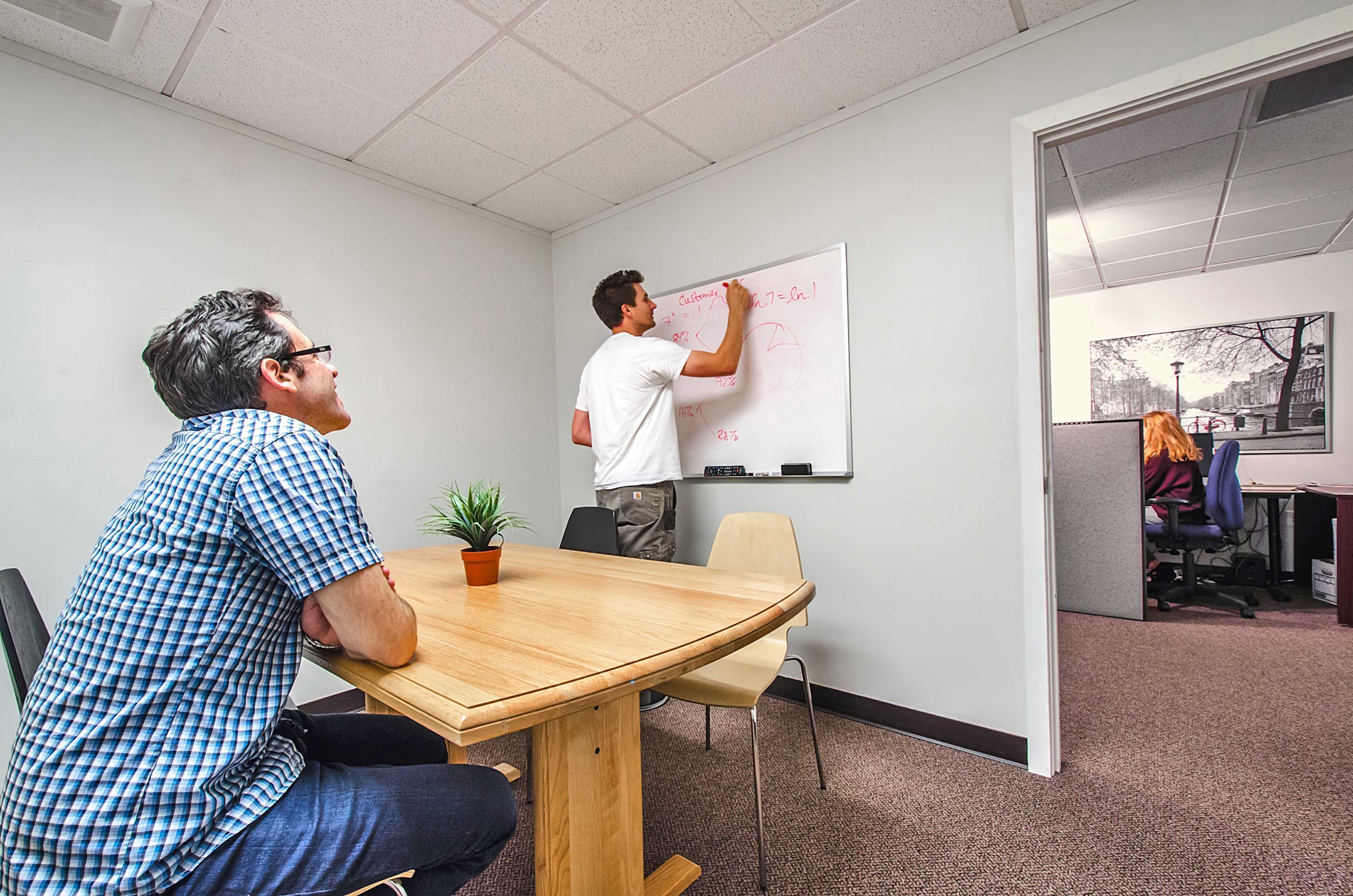 A man writes on a whiteboard while another man observes, seated at a wooden table in a small office meeting room.