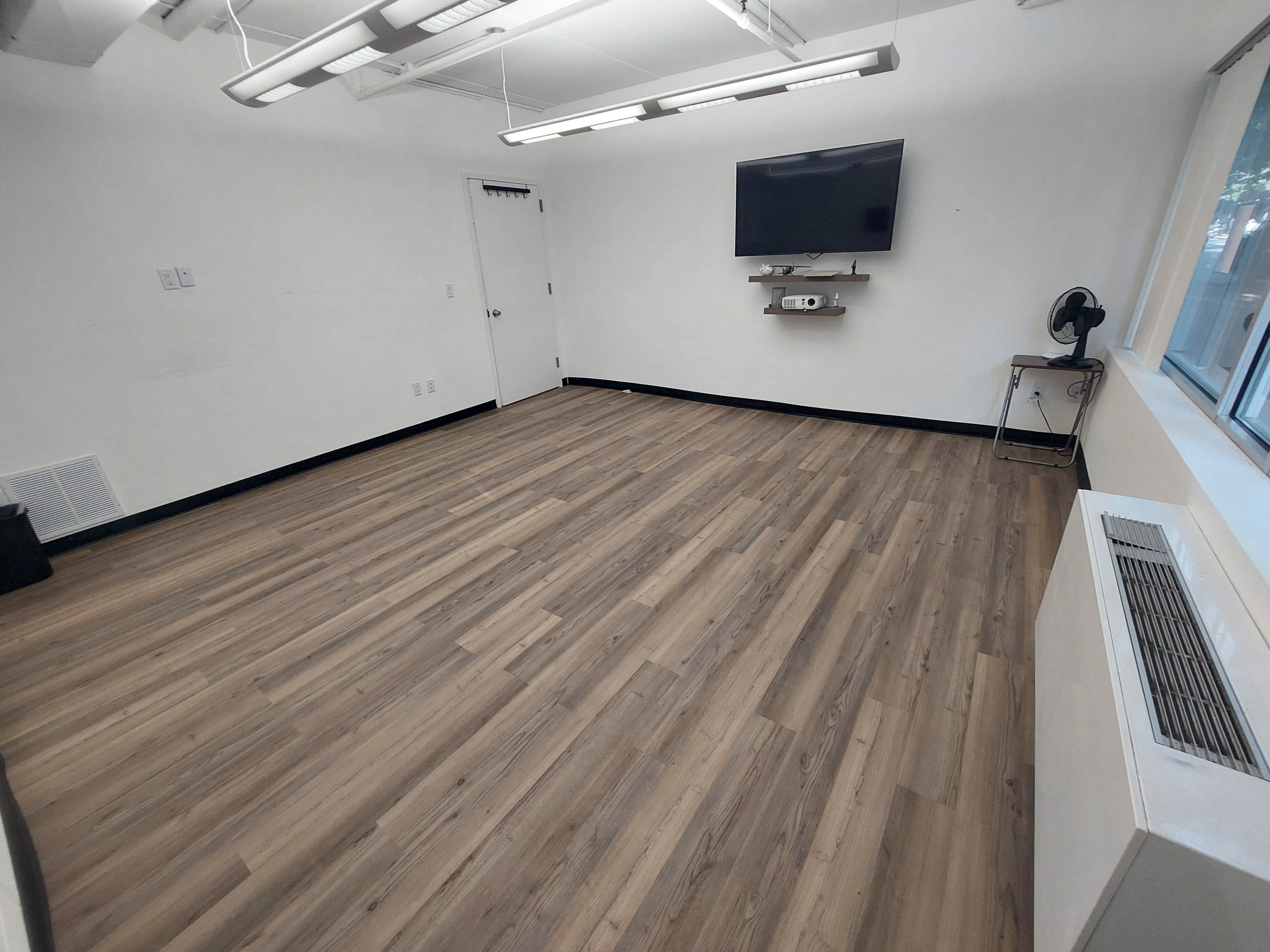 The image shows an empty room with wooden flooring, a television mounted on the wall, a fan on a small table, and large windows letting in natural light.