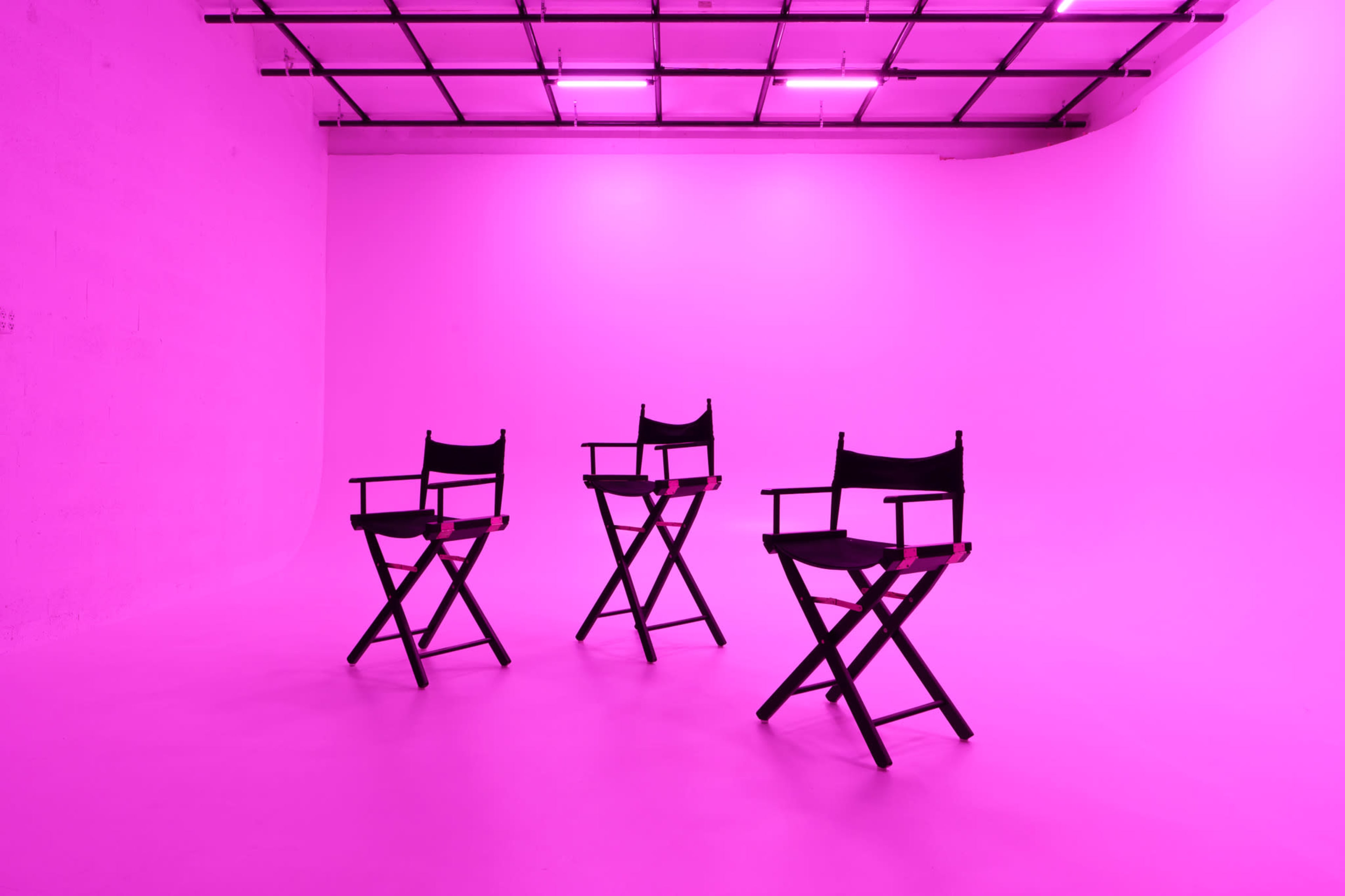 Three black director's chairs are positioned in a brightly lit, pink studio space with a smooth floor.