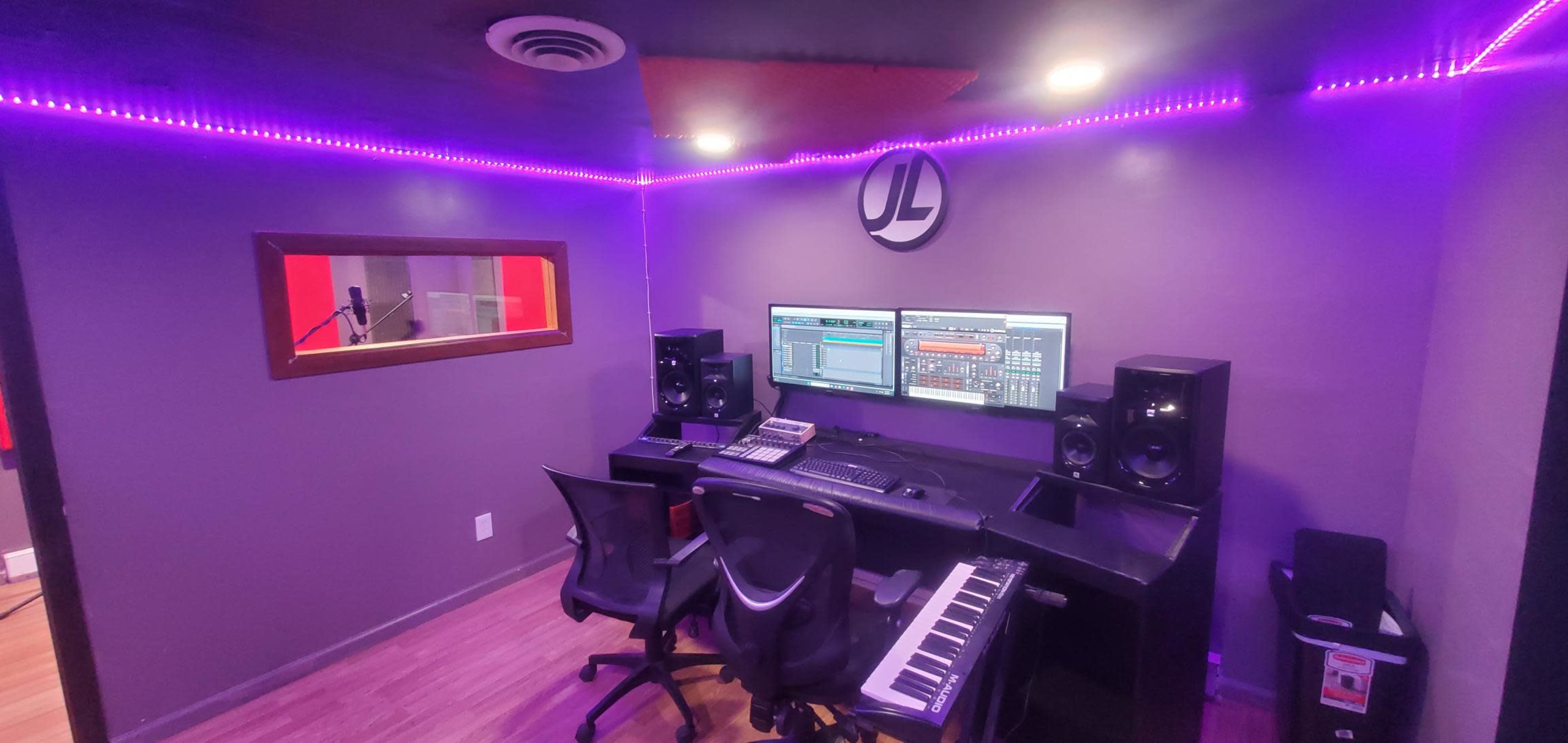 A modern music production studio featuring a central workspace with two computer monitors, audio equipment, and a MIDI keyboard, illuminated by purple LED lighting.
