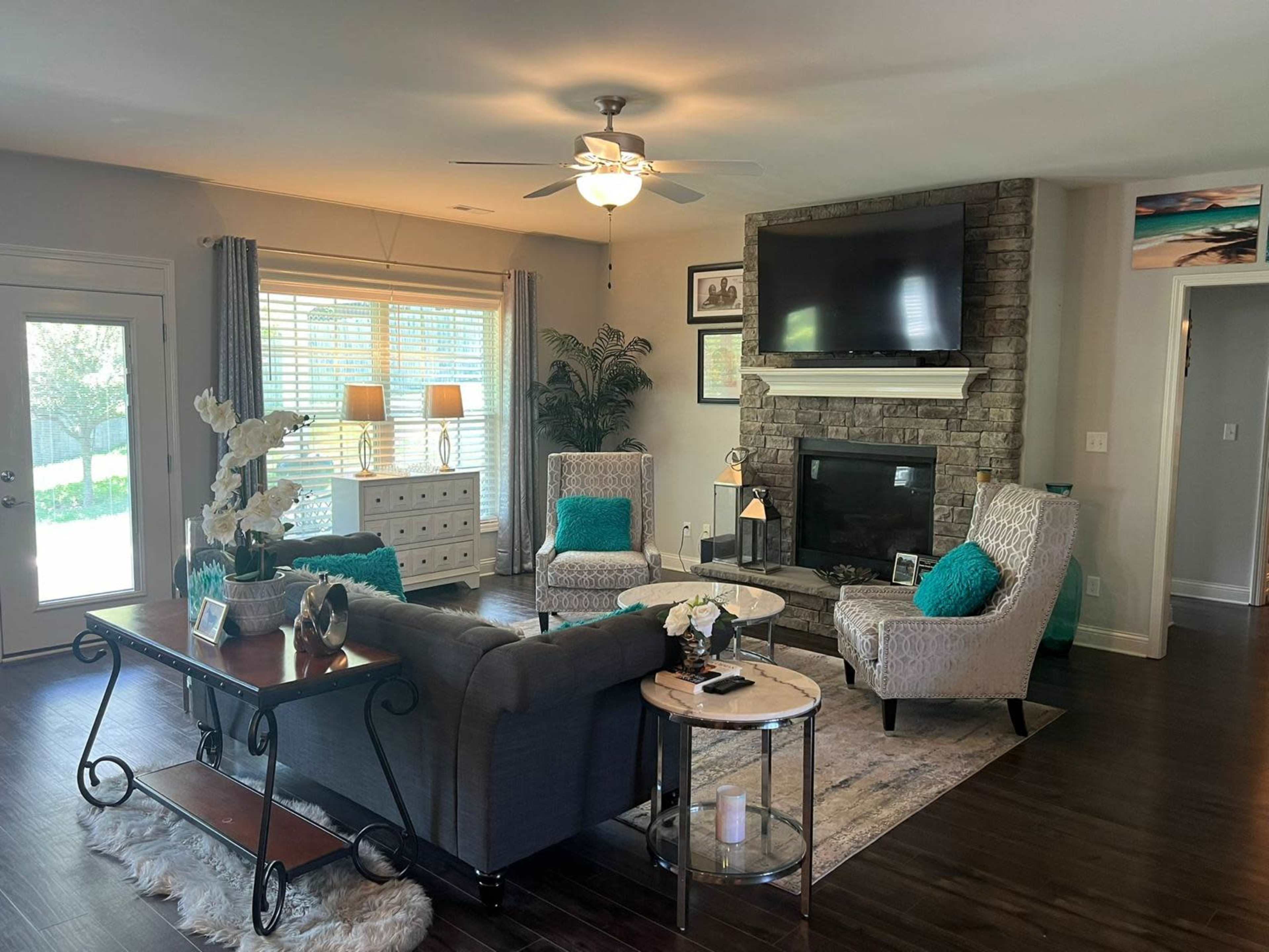 The living room features a gray sofa and two armchairs with teal accents, a stone fireplace, and large windows allowing natural light.