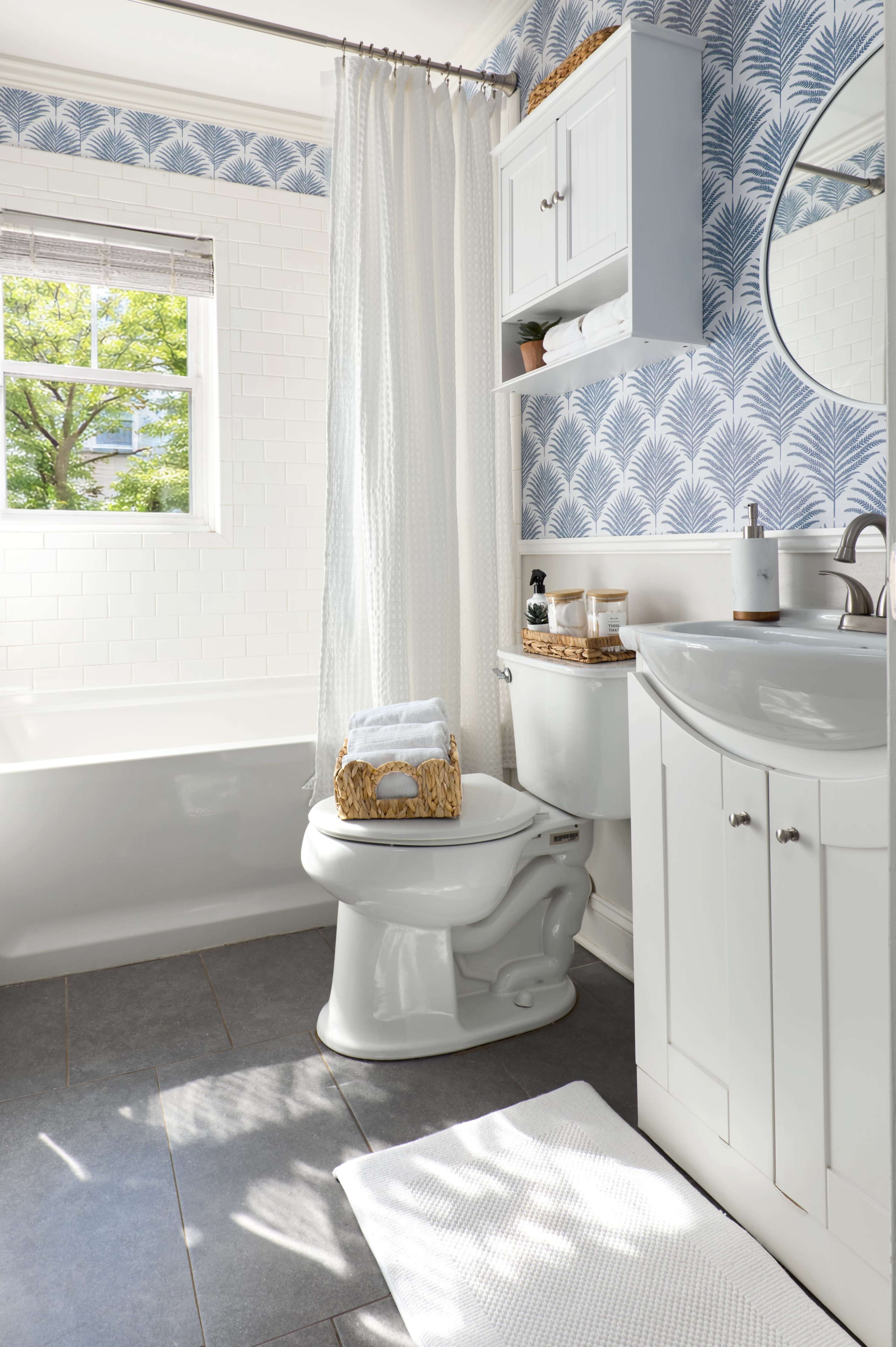The bathroom features a white tub, a round mirror above the sink, and blue patterned wallpaper, with a towel and decorative items arranged on the toilet.