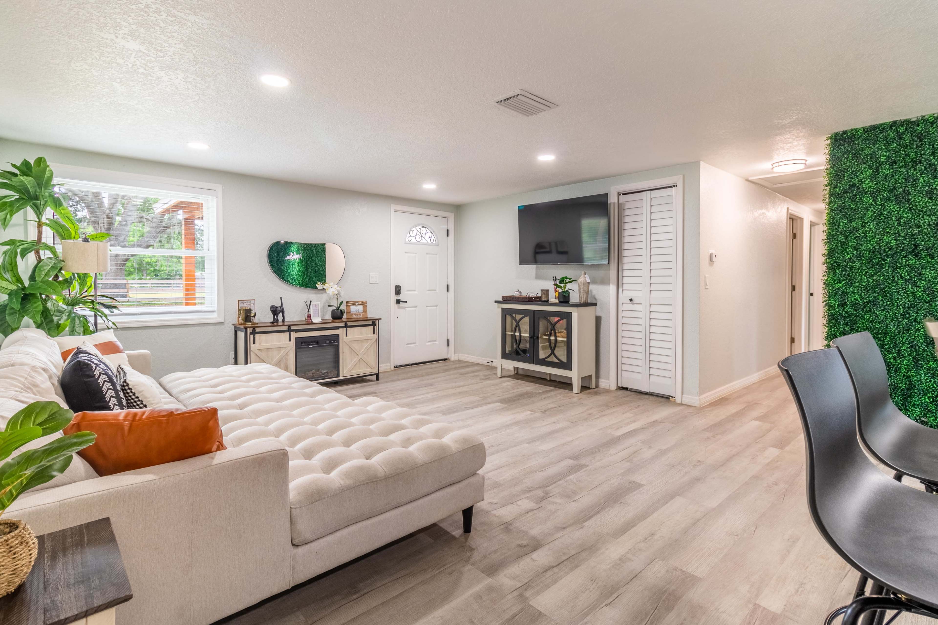 A modern living room with a beige couch, a flat-screen TV on the wall, a small kitchenette area, and green accents from plants and wall decor.