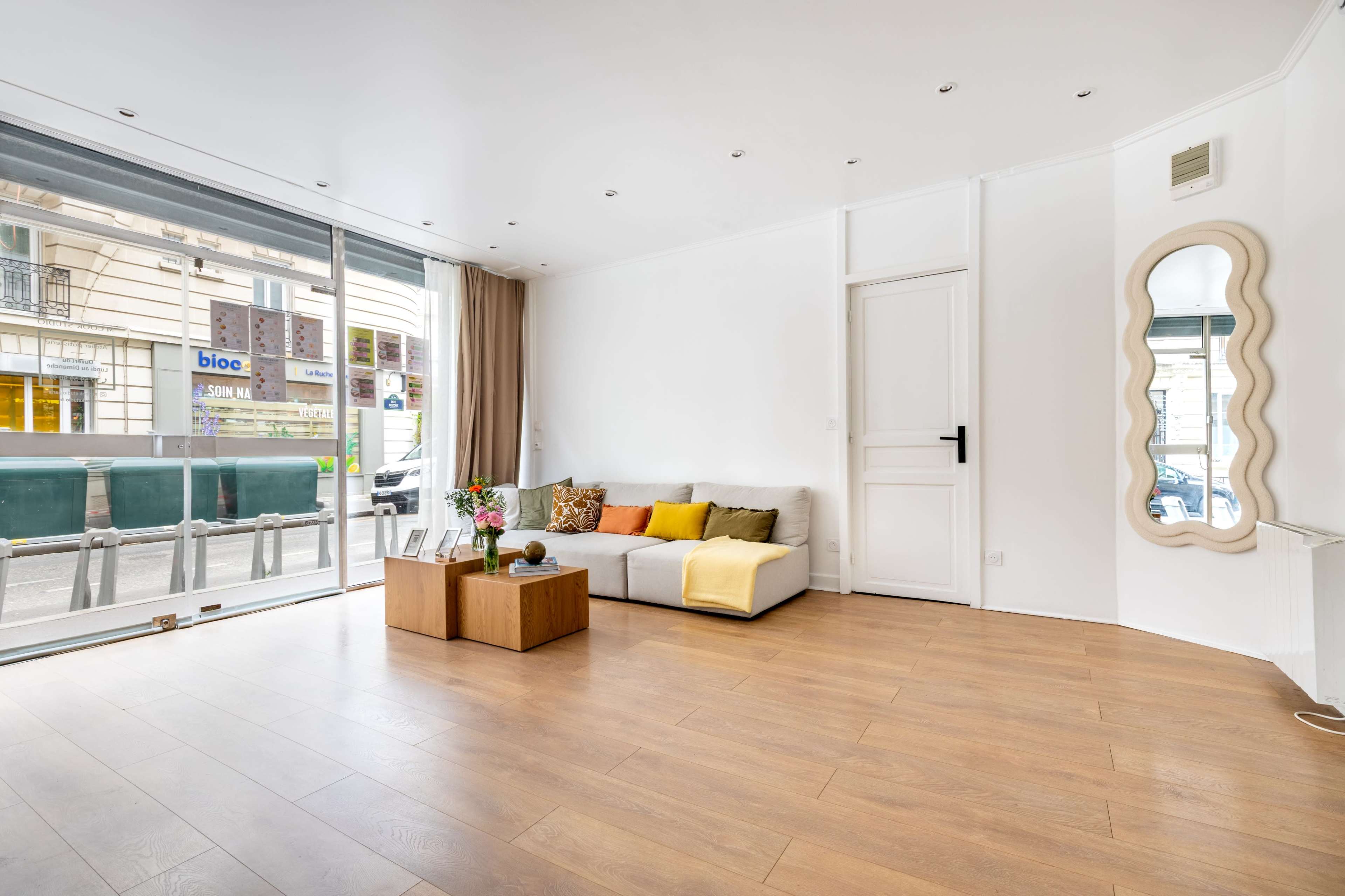 A bright living room features a gray sofa with colorful pillows, a coffee table, and large windows that allow natural light to fill the space.