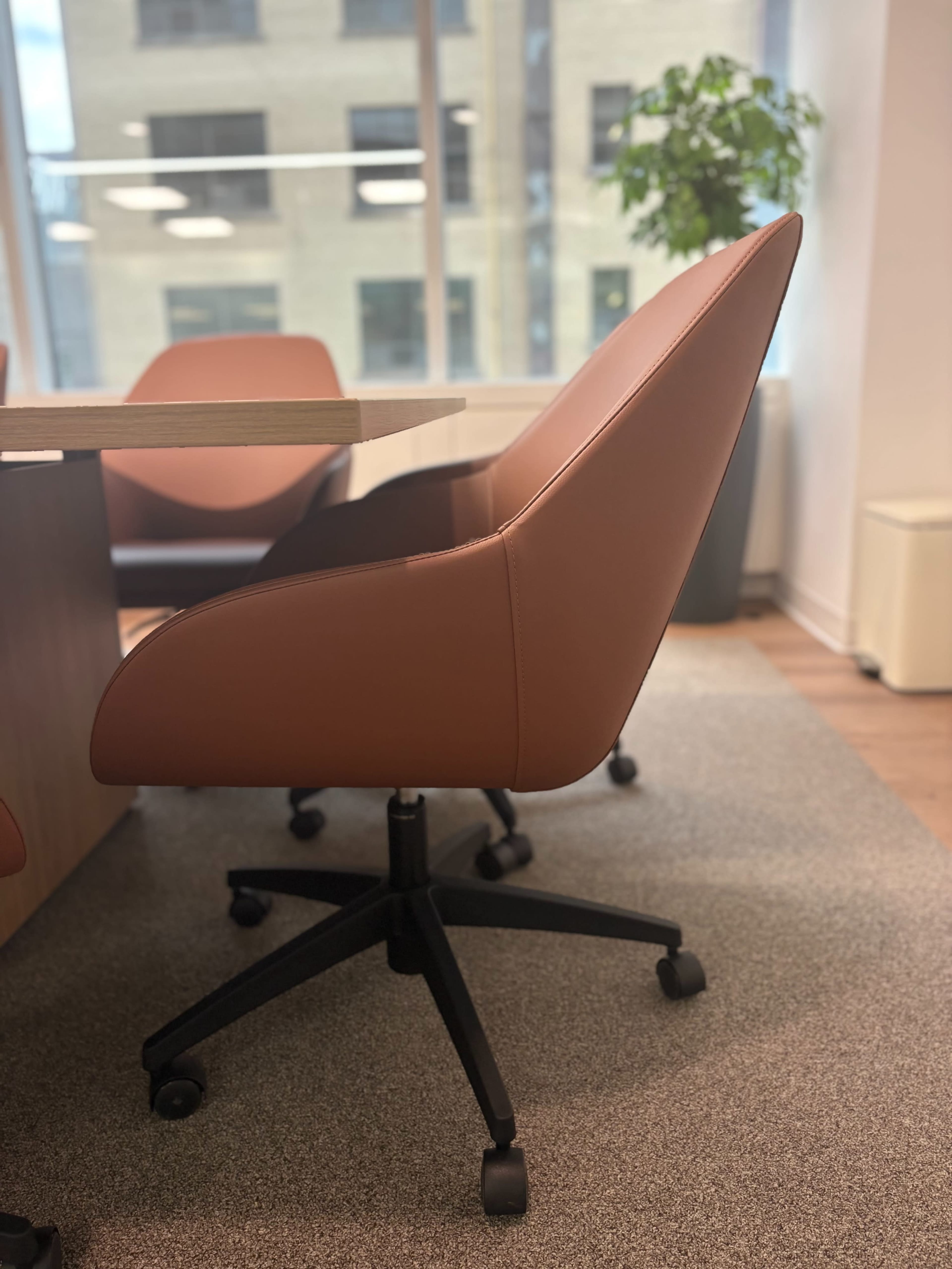A modern office chair with a brown leather finish sits beside a wooden conference table in a well-lit room.