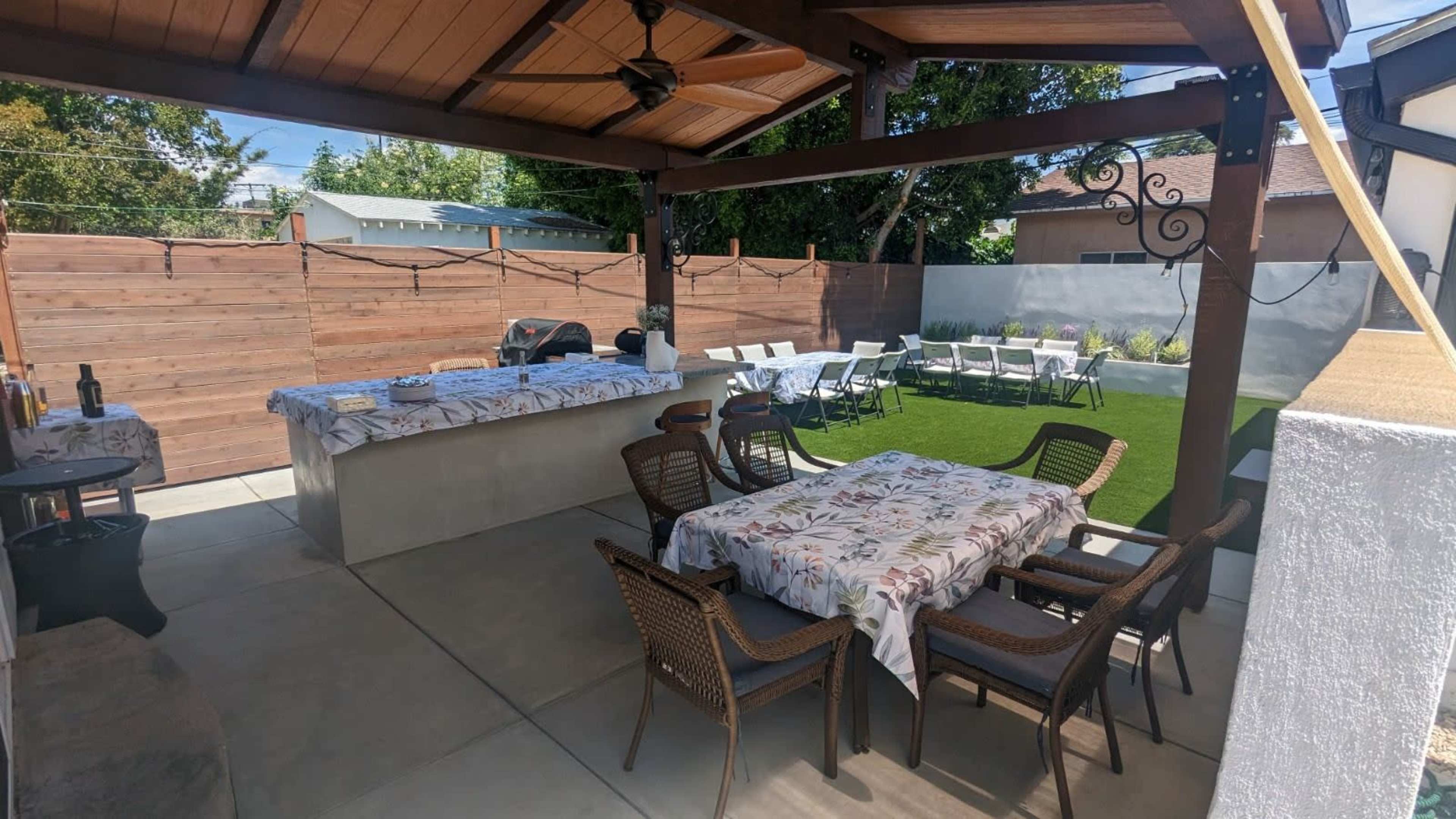 A covered outdoor patio features a table with floral tablecloths, wicker chairs, and an adjacent area with additional seating and a barbecue setup.