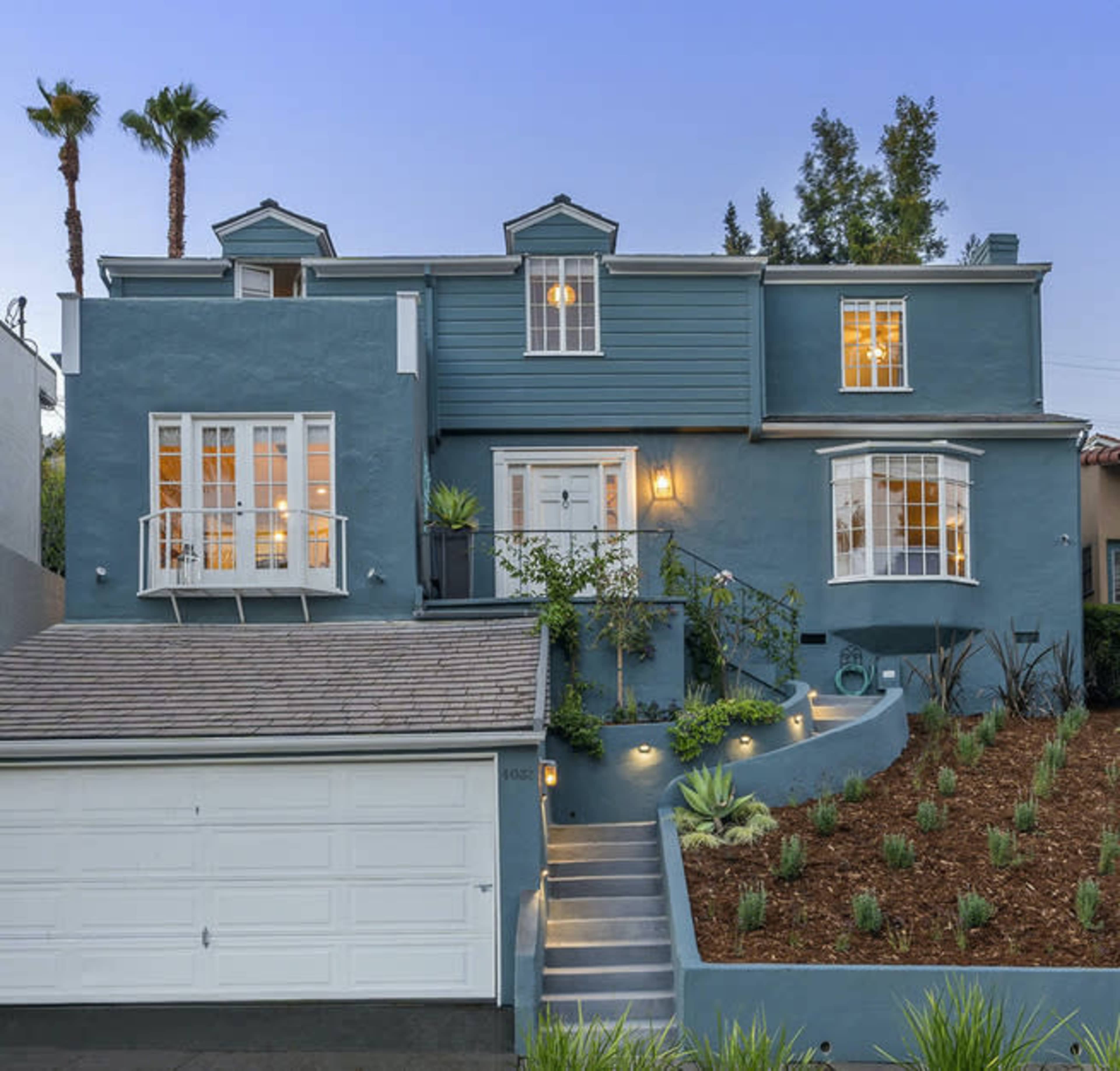 A two-story blue house with multiple windows and a landscaped front yard is set on a sloped driveway.