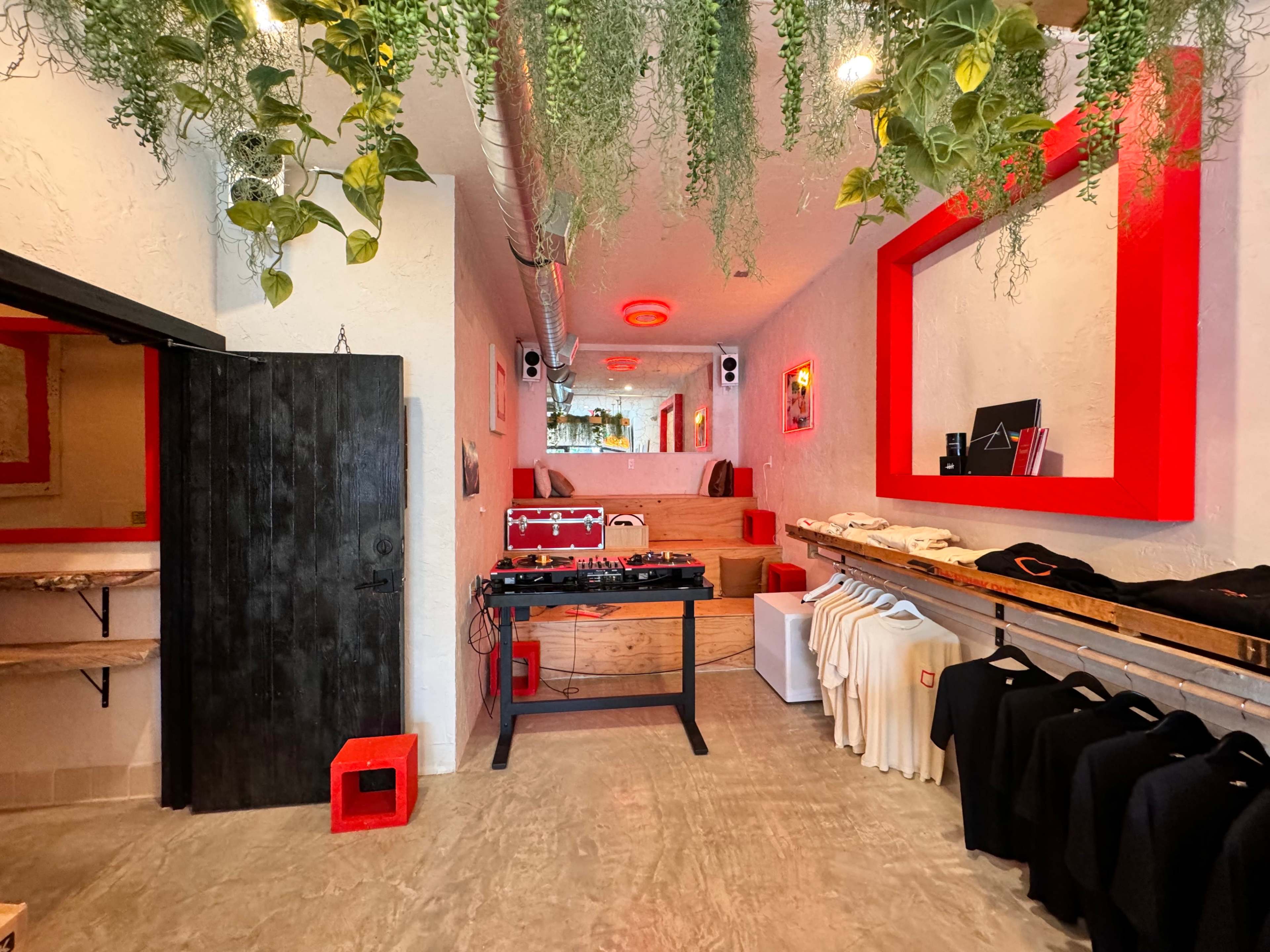 An interior of a boutique with a display of clothing items, a DJ setup in the center, and greenery hanging from the ceiling.