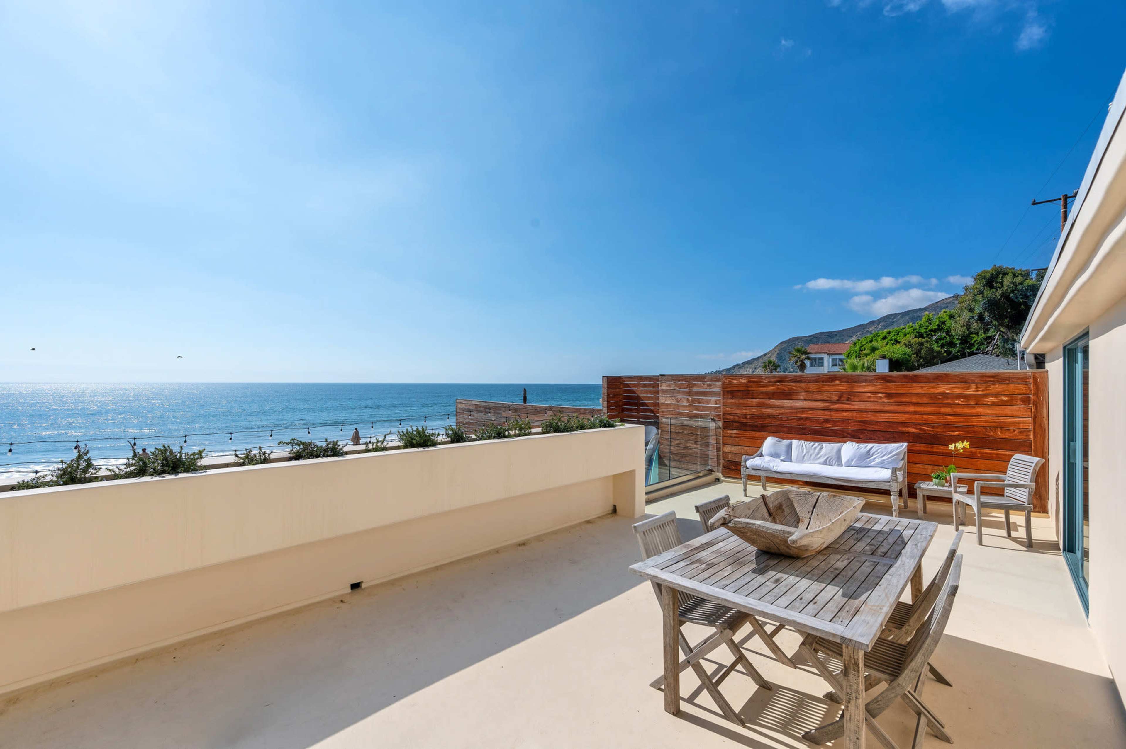 A patio with a wooden dining table and chairs overlooking a beach and ocean under a clear blue sky.