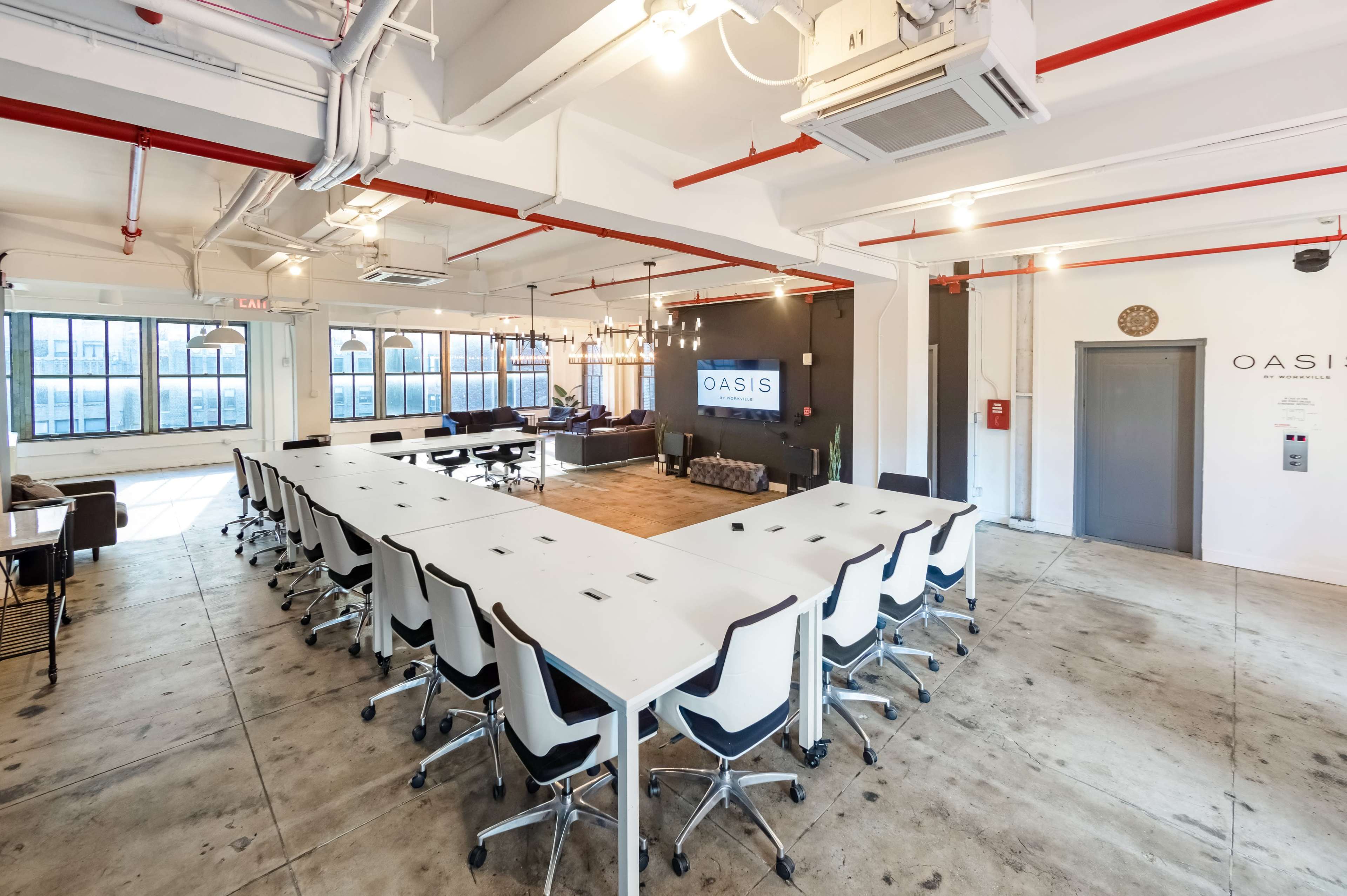 A modern office space features a large, U-shaped table surrounded by chairs, with large windows and a lounge area in the background.