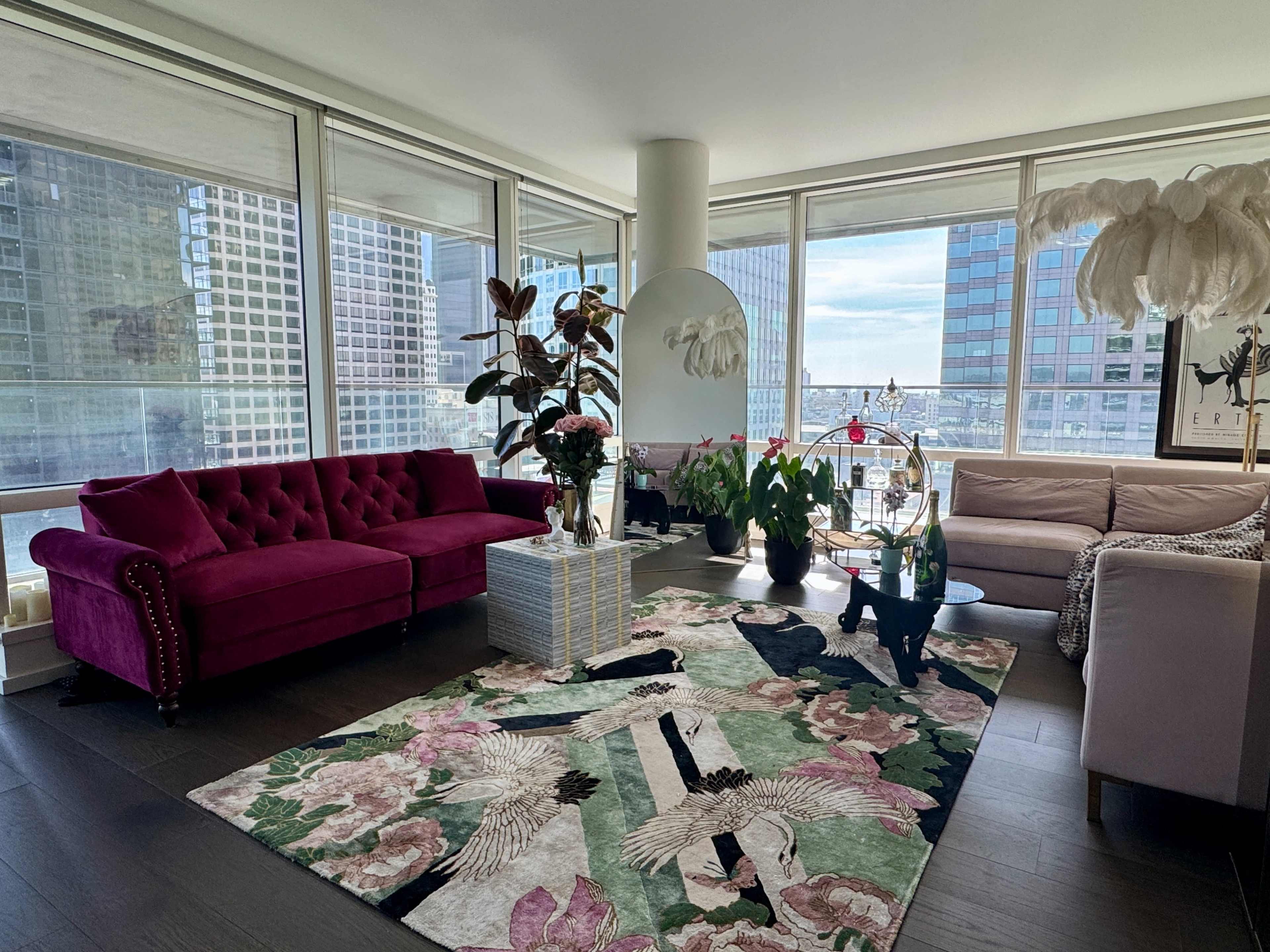 The image shows a modern living room featuring two distinct sofas, a large area rug with floral and bird motifs, and several indoor plants, all illuminated by natural light from large windows.