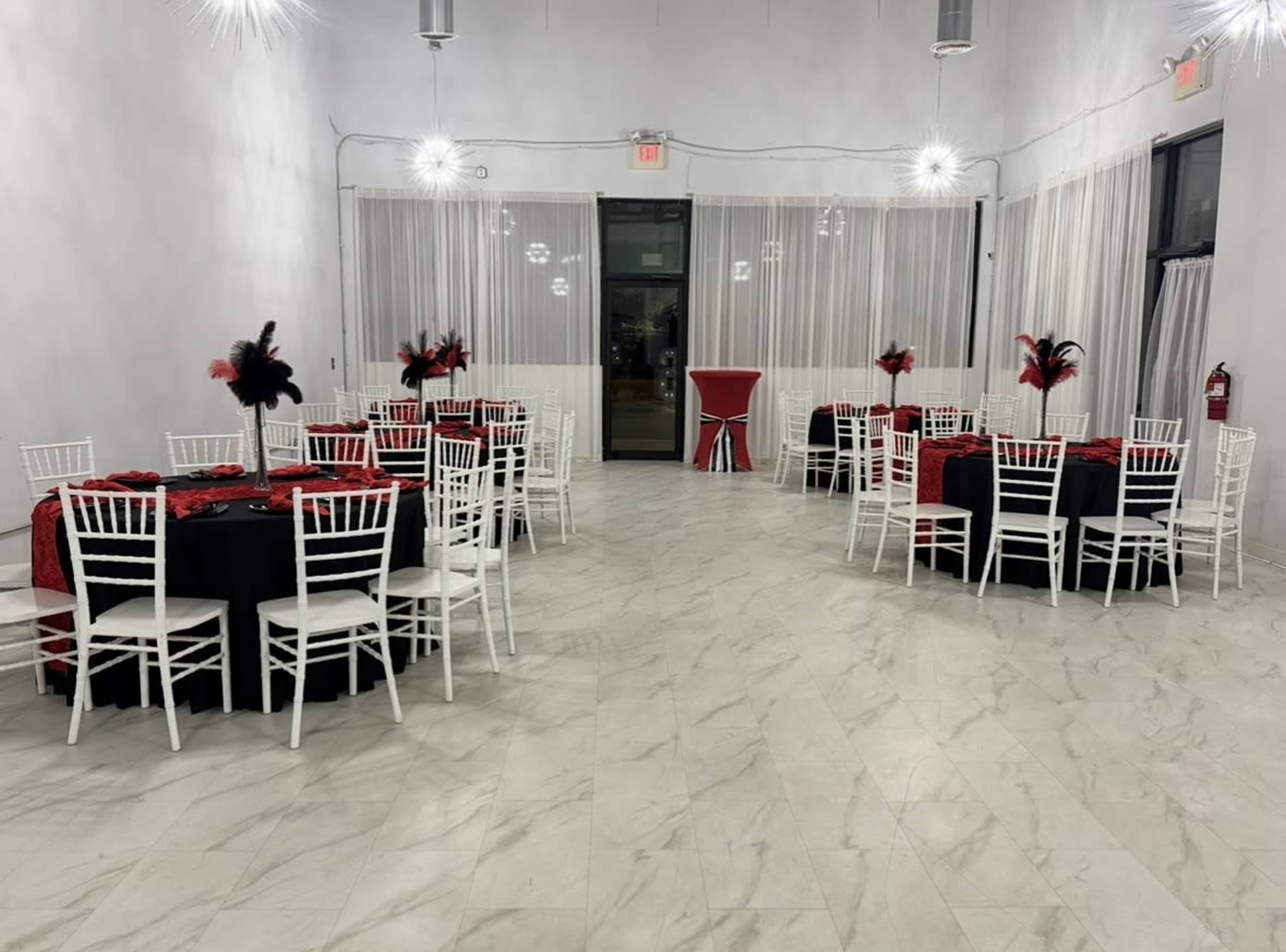 The image shows a modern event space set up with round tables covered in black and red tablecloths, surrounded by white chairs and adorned with feather centerpieces.