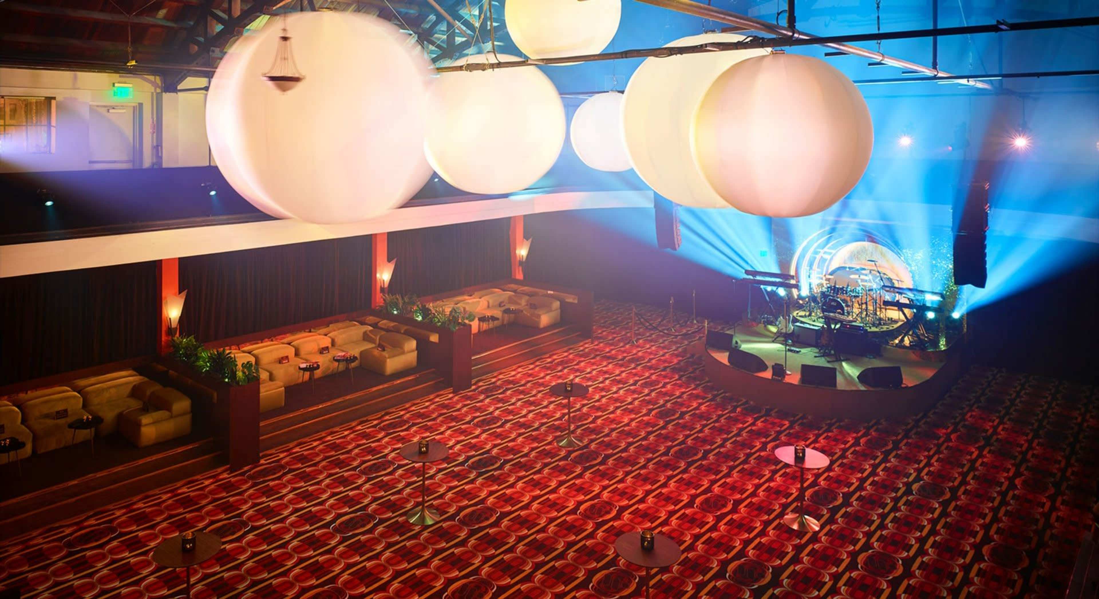 The image shows a spacious indoor venue with a stage set up for a performance, featuring a patterned carpet and large hanging spherical lights.