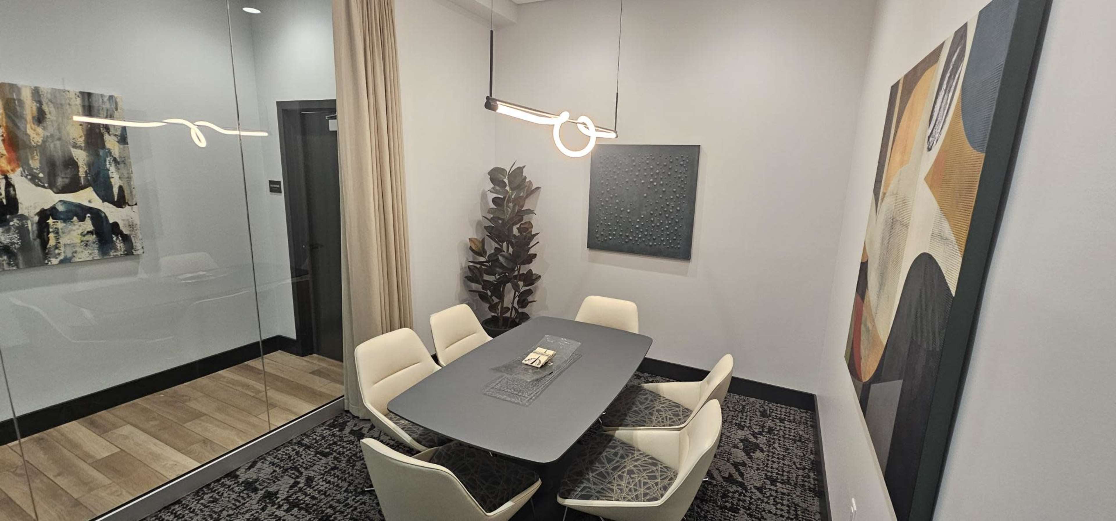 The image shows a modern meeting room with a rectangular table surrounded by white chairs, illuminated by a hanging light fixture, and adorned with abstract art on the walls.