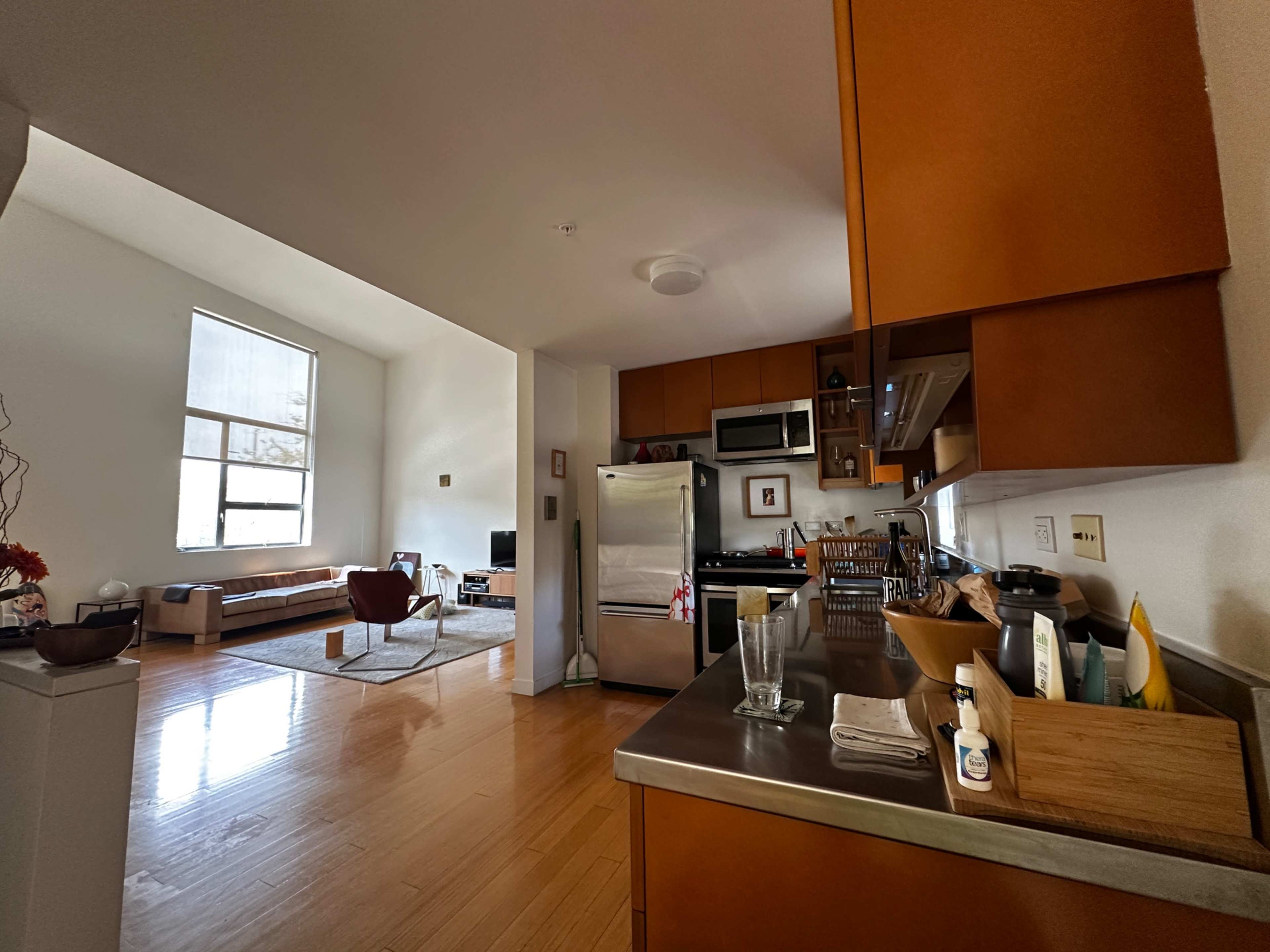 The image shows a modern kitchen with brown cabinetry and stainless steel appliances, connected to a living area that features a sofa and a large window.