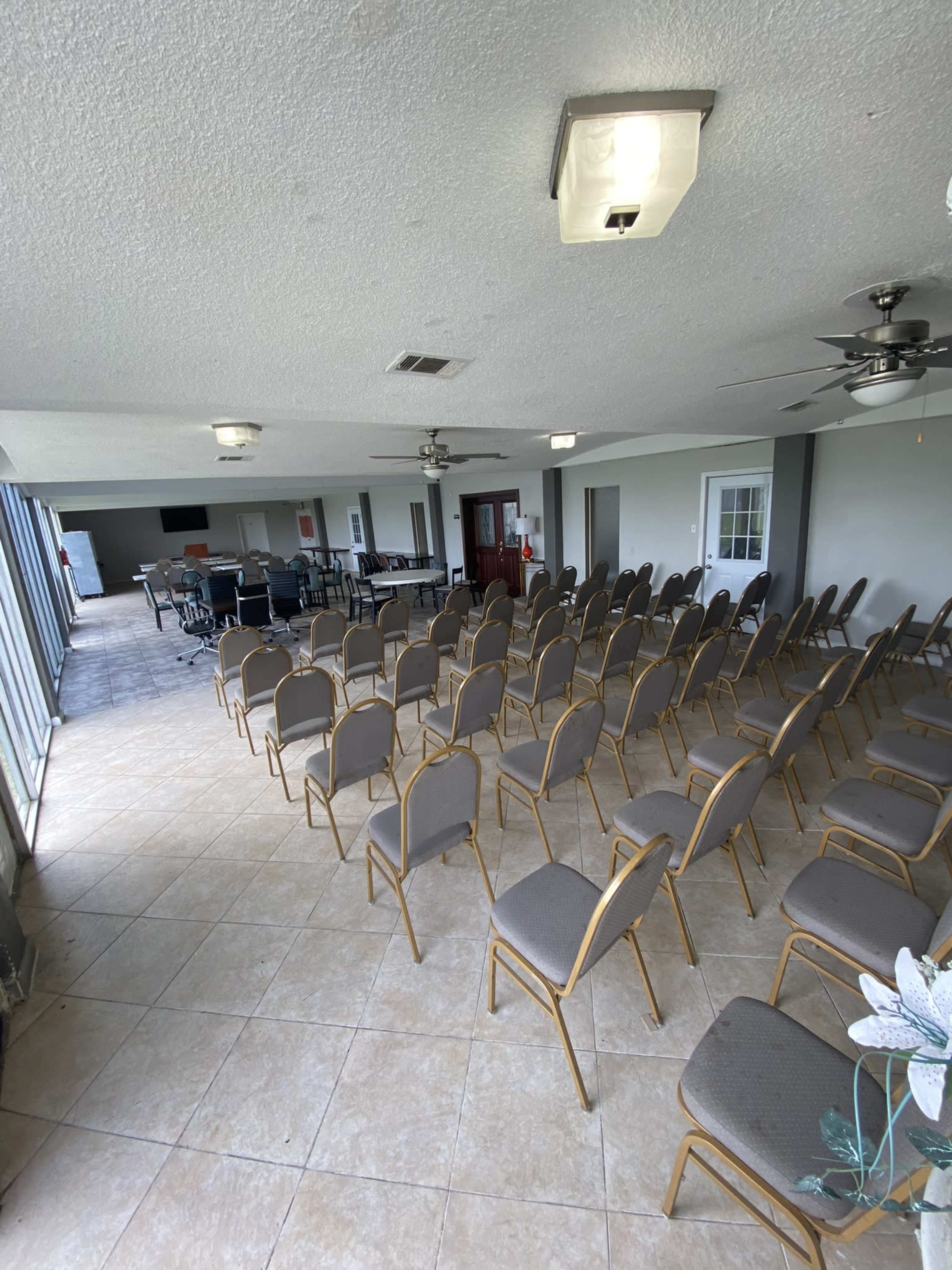 A spacious room arranged with multiple rows of empty chairs, featuring ceiling fans and large windows.