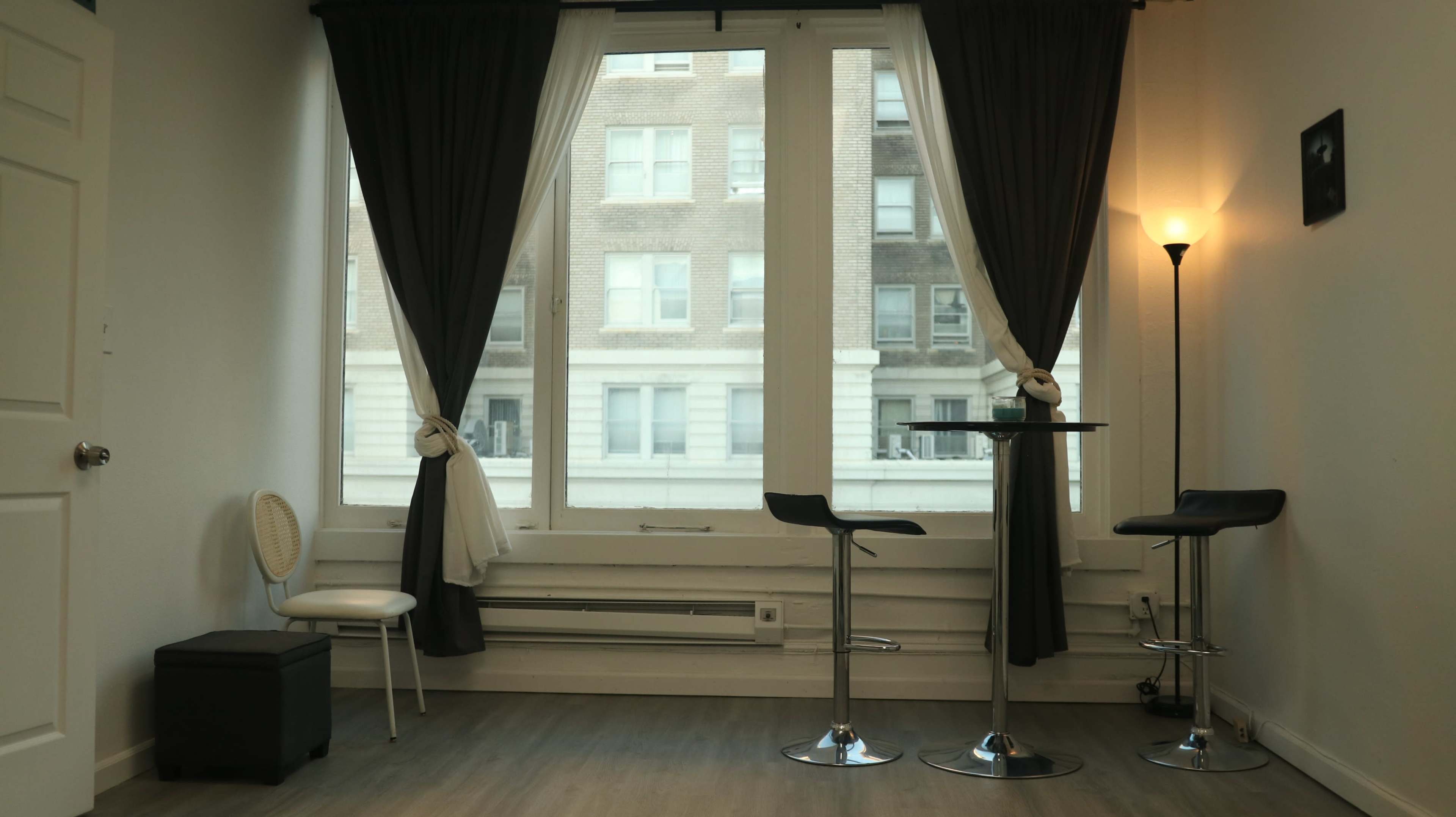 The image shows a modern interior space with large windows, bar stools, and a small table, framed by dark curtains.