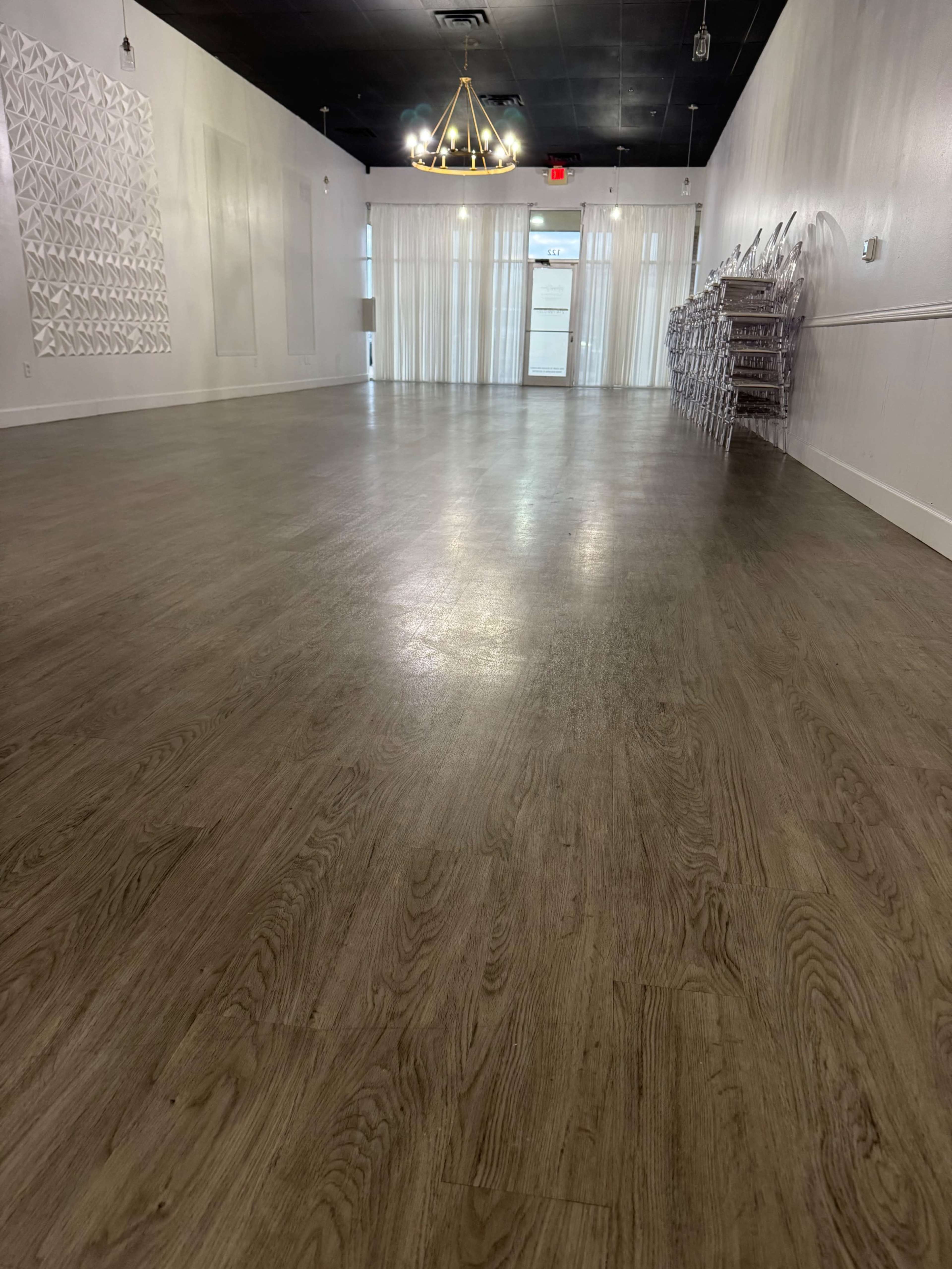 The image shows an empty indoor space with wooden flooring, large windows, and white walls.