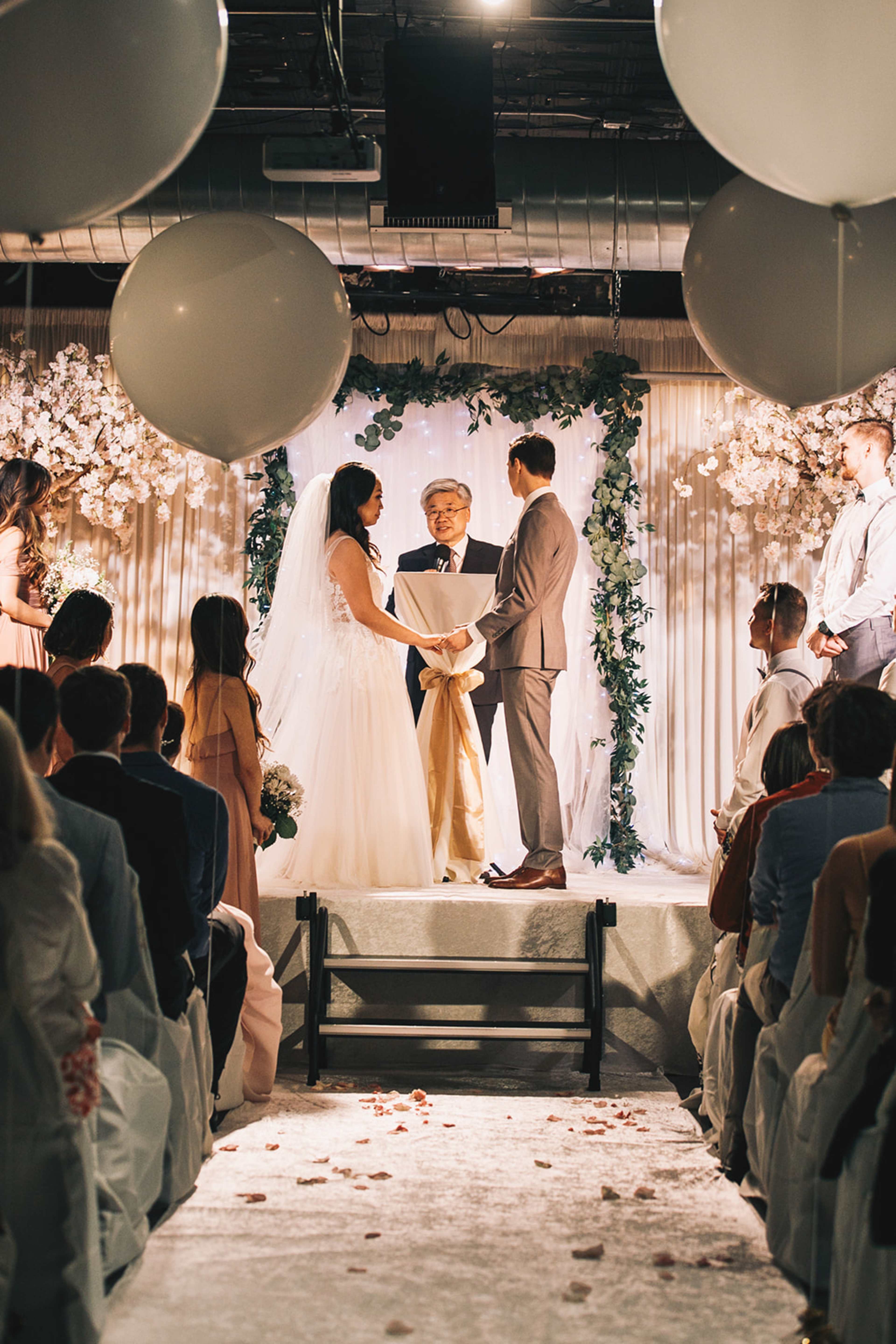 A couple stands at an altar decorated with flowers, exchanging vows in front of an officiant, while guests seated on either side watch.