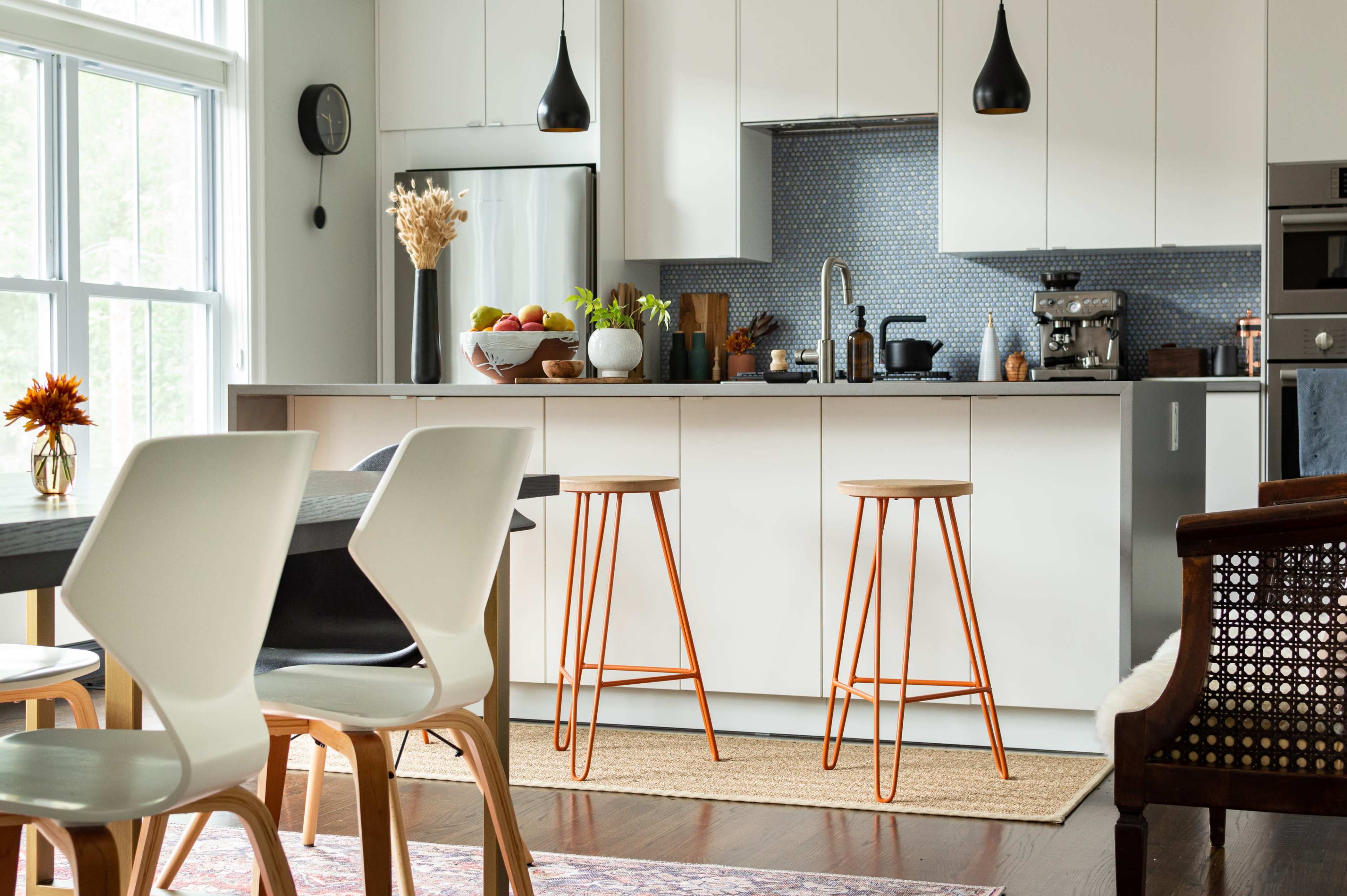 A modern kitchen features white cabinetry, a blue tiled backsplash, two metal bar stools with wooden tops, and a dining area with white chairs.