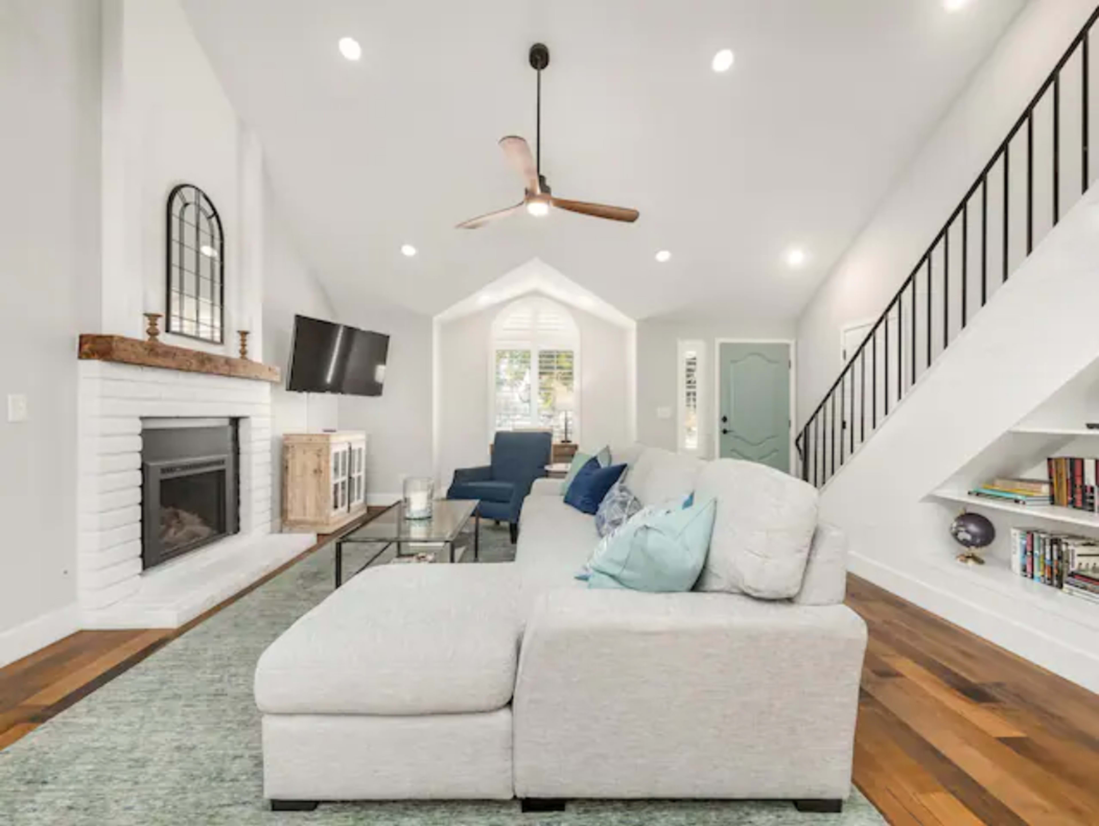 A light-filled living room features a sectional sofa, a fireplace, and a staircase leading to an upper level.