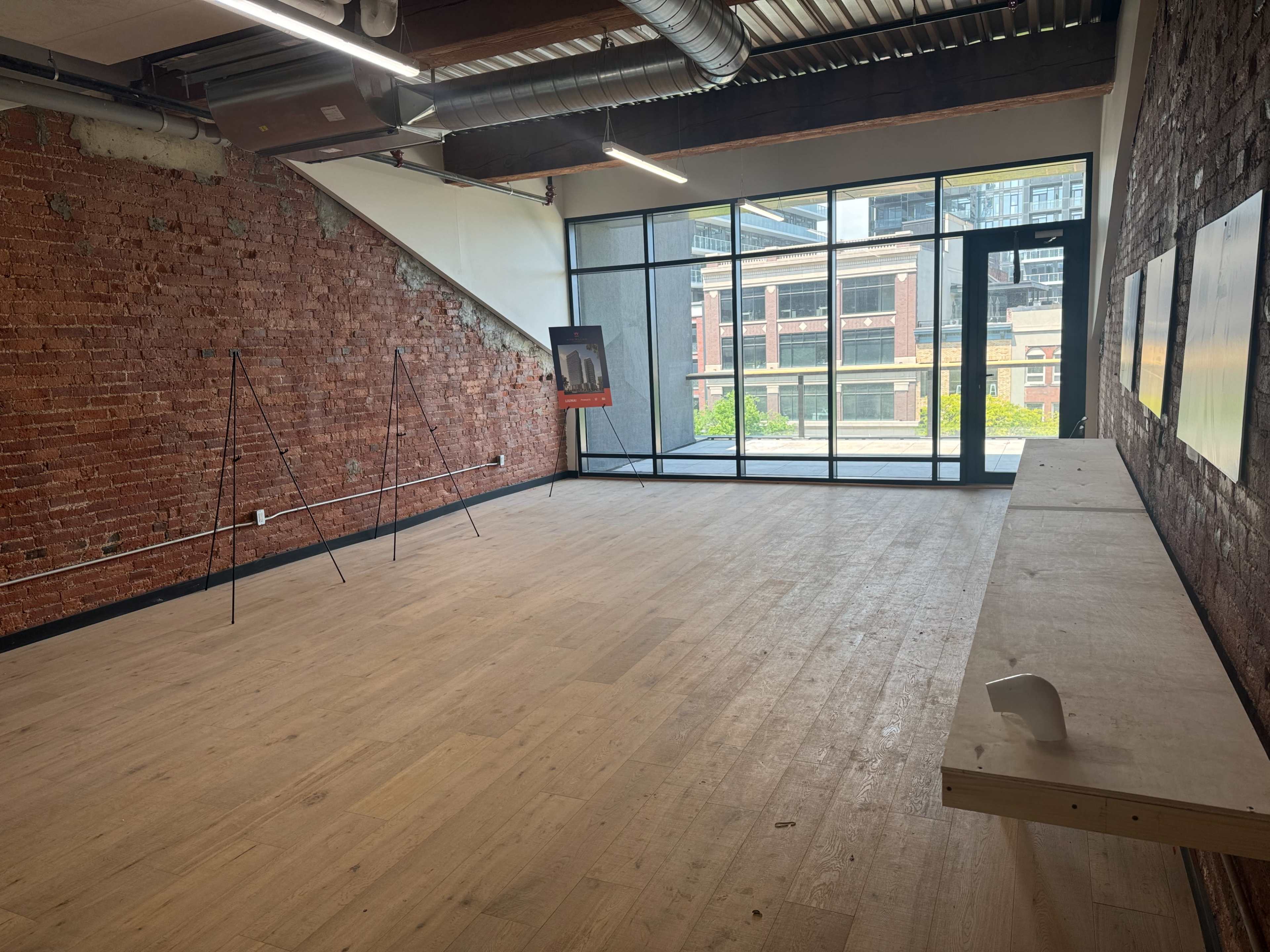 An empty room with exposed brick walls, large windows, and light wooden flooring, featuring a simple wooden shelf against one wall.