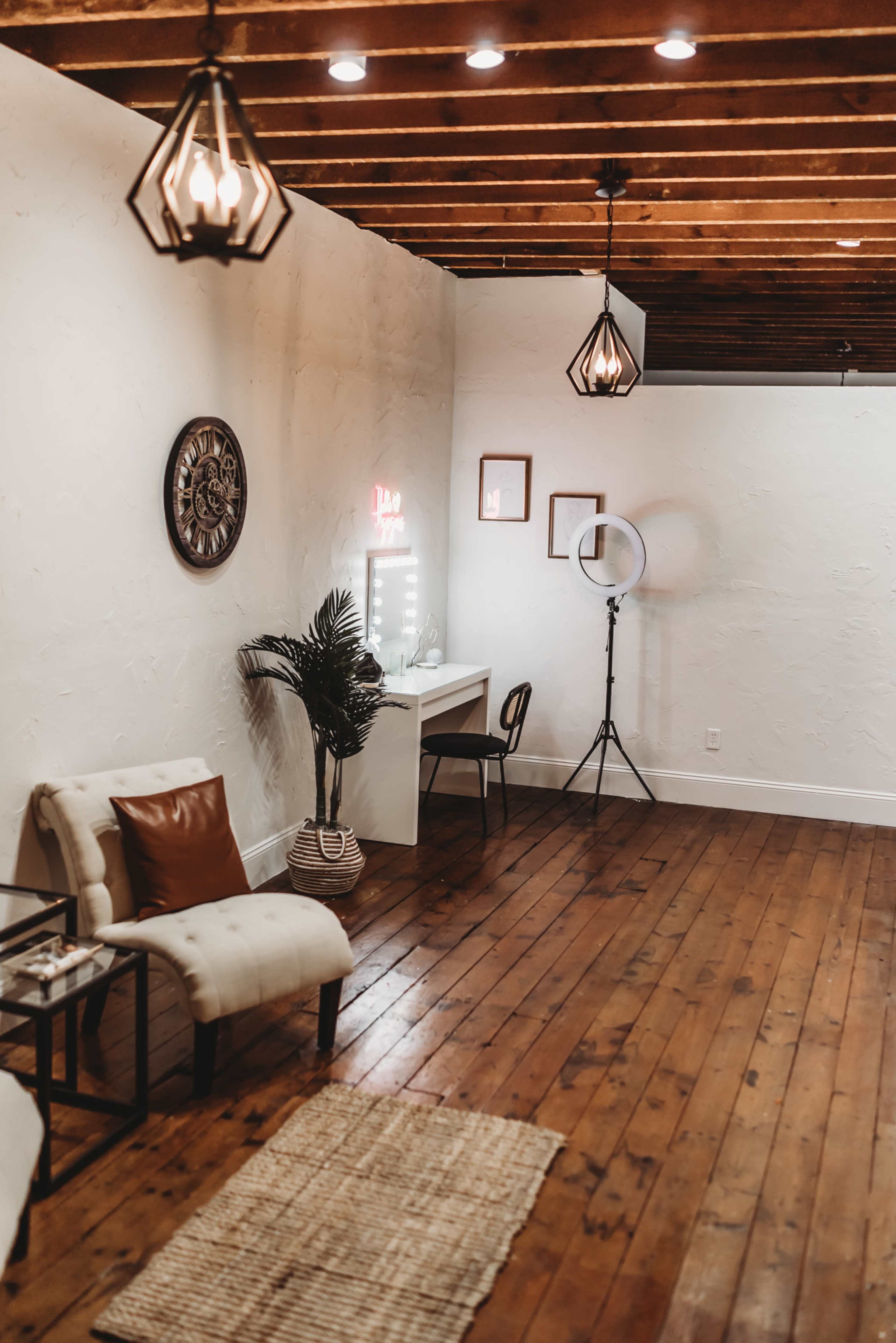 The image shows a well-lit room with wooden flooring, featuring a desk with a mirror, a comfortable chair, decorative lighting, and a potted plant.