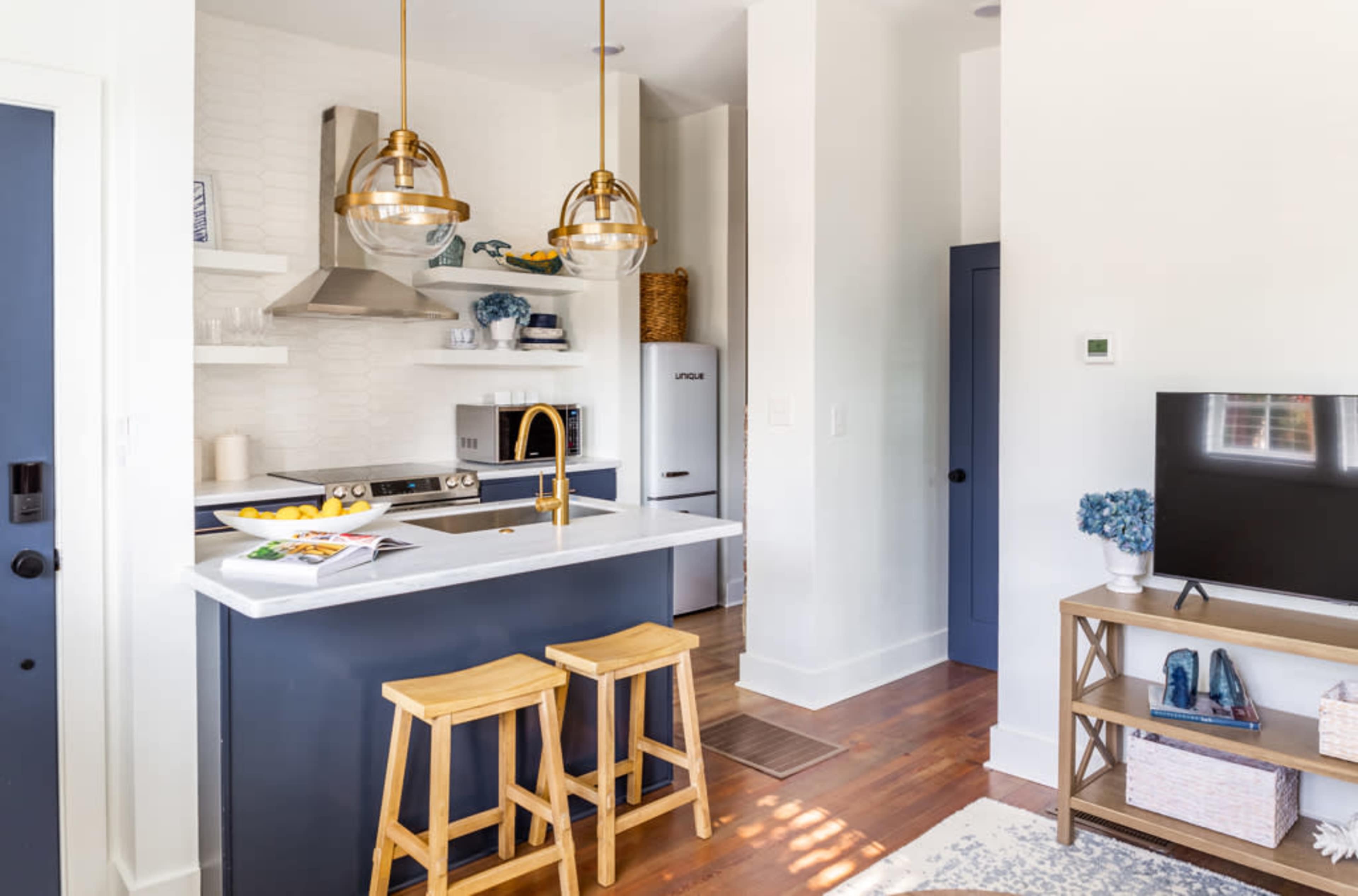 A modern kitchenette features a white countertop, gold pendant lights, and wooden bar stools, with an open layout leading to a living area.