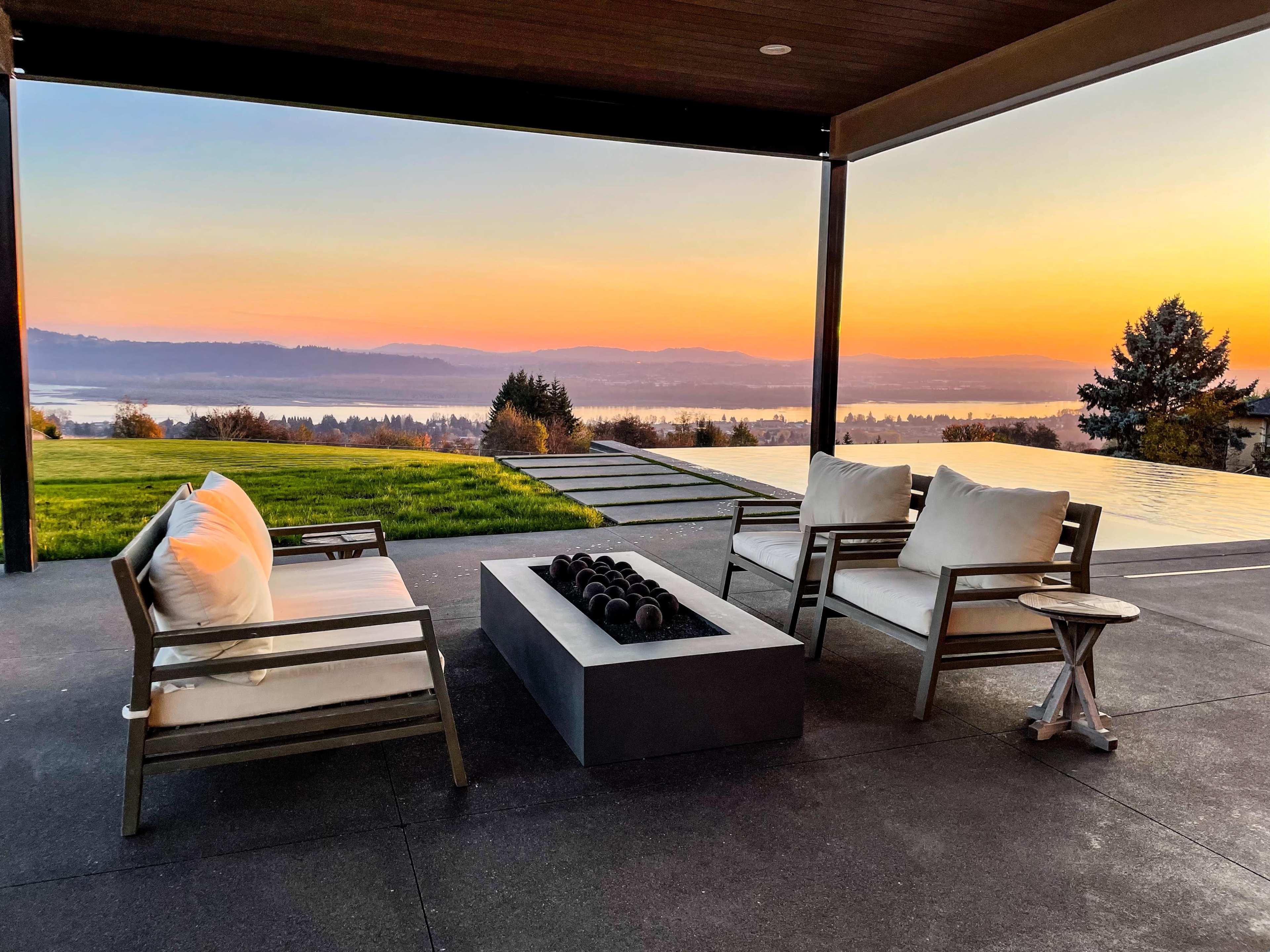 A patio area features two chairs and a fire pit overlooking a lake at sunset.