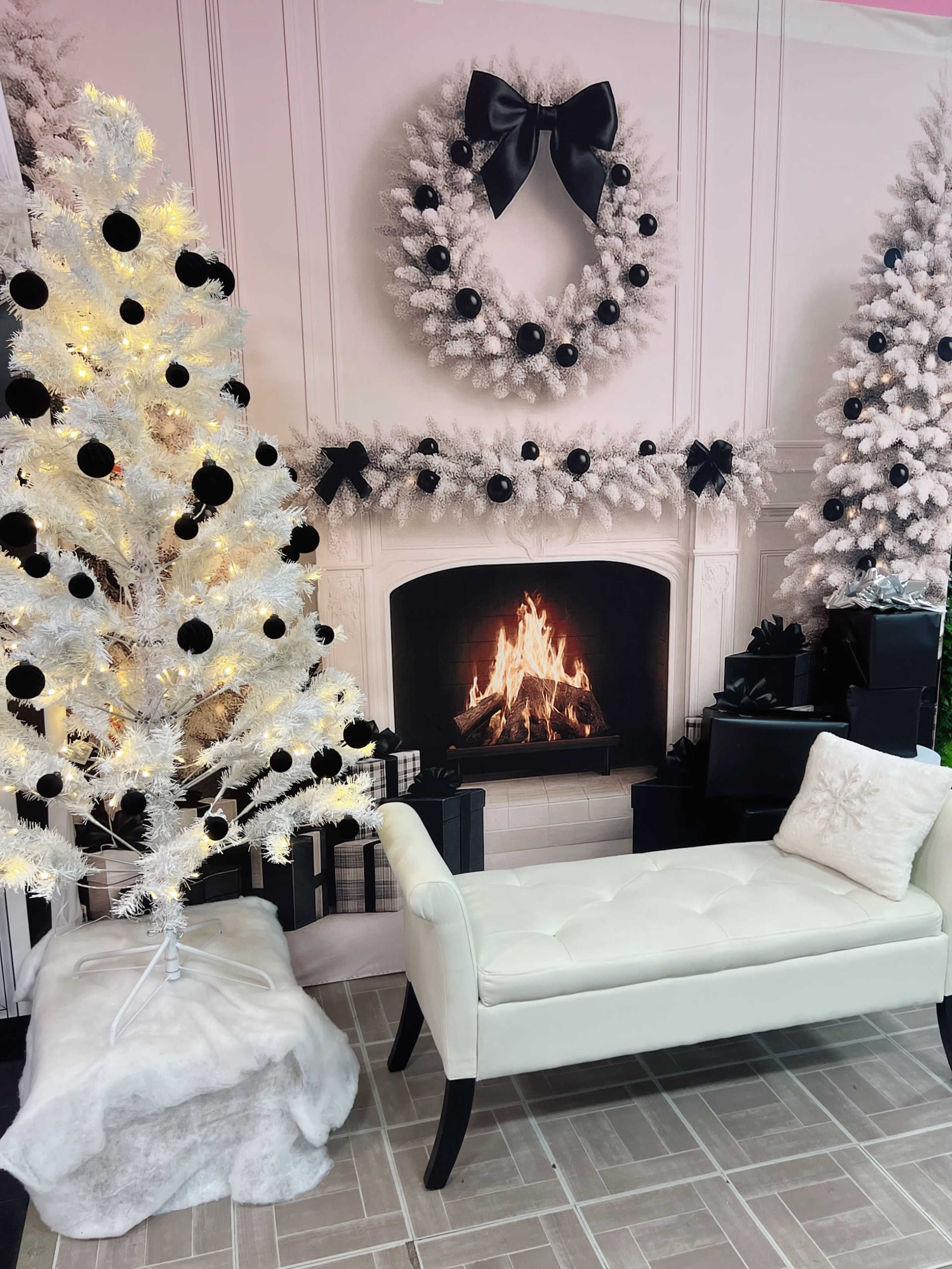 The scene features a white tree decorated with black ornaments next to a fireplace with a lit fire, surrounded by a backdrop of a festive wreath and holiday decorations.