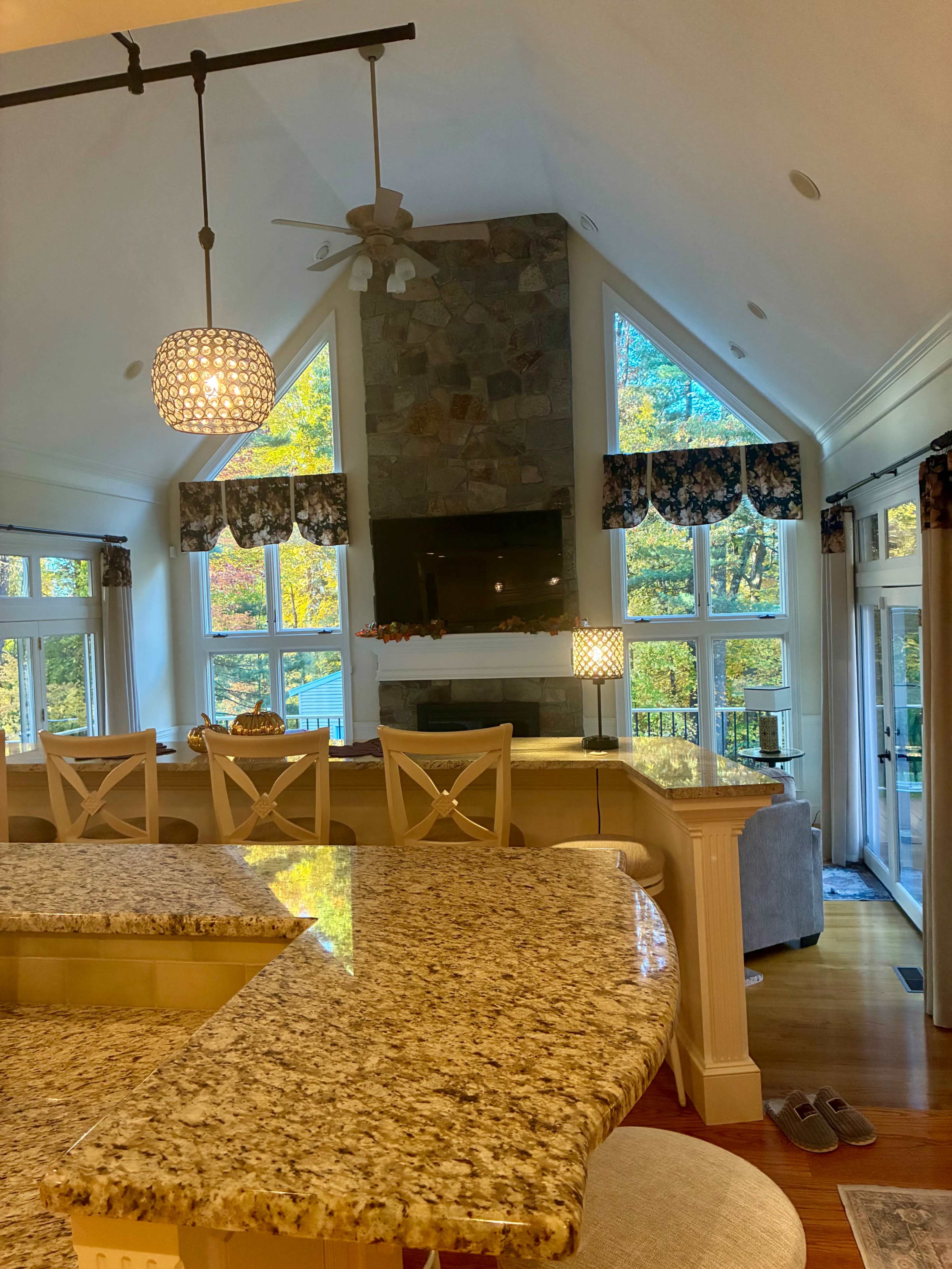 The image shows a cozy living area with a stone fireplace, large windows allowing natural light, and a kitchen bar with granite countertops.