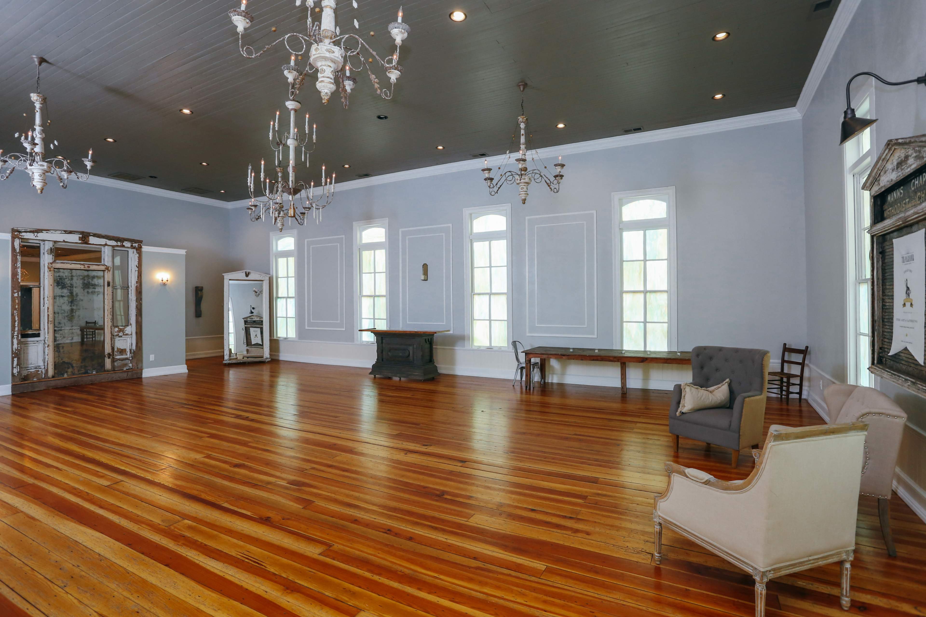 A spacious room with hardwood flooring, large windows, ornate chandeliers, and minimal furniture, including a couple of chairs and a wooden table.