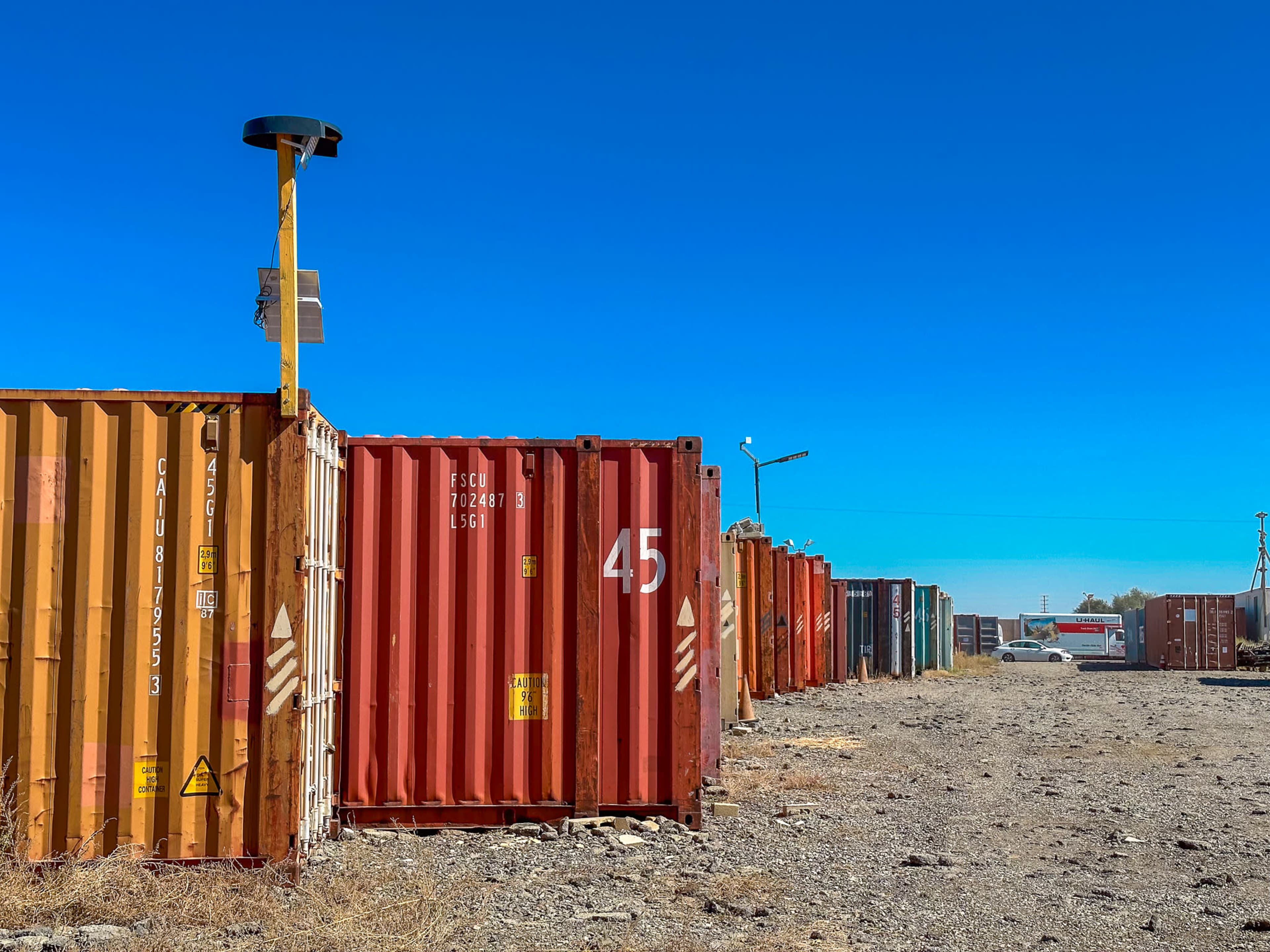 A row of multi-colored shipping containers stands in an open area under a clear blue sky.