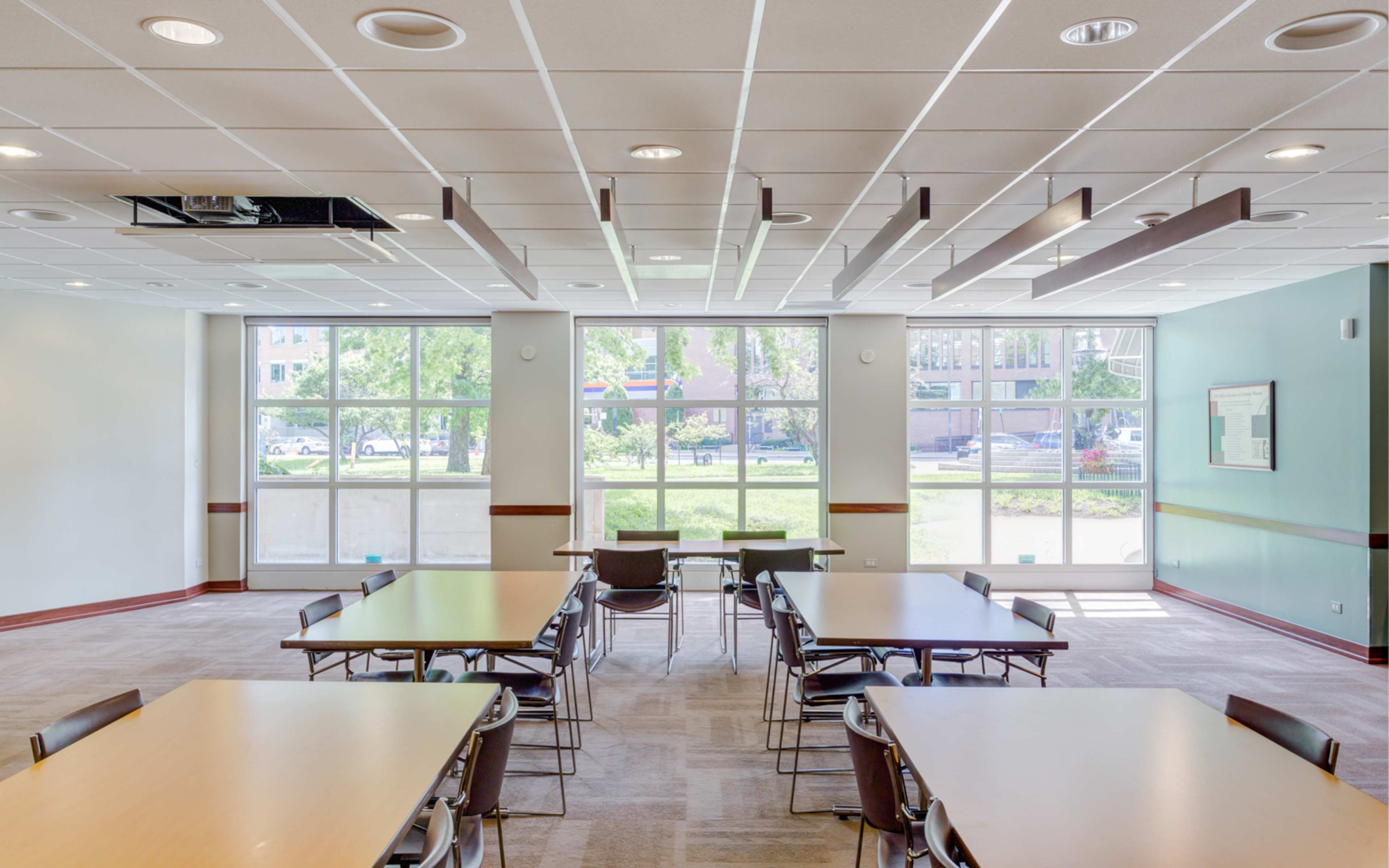 A spacious room with several tables and chairs, illuminated by natural light from large windows.