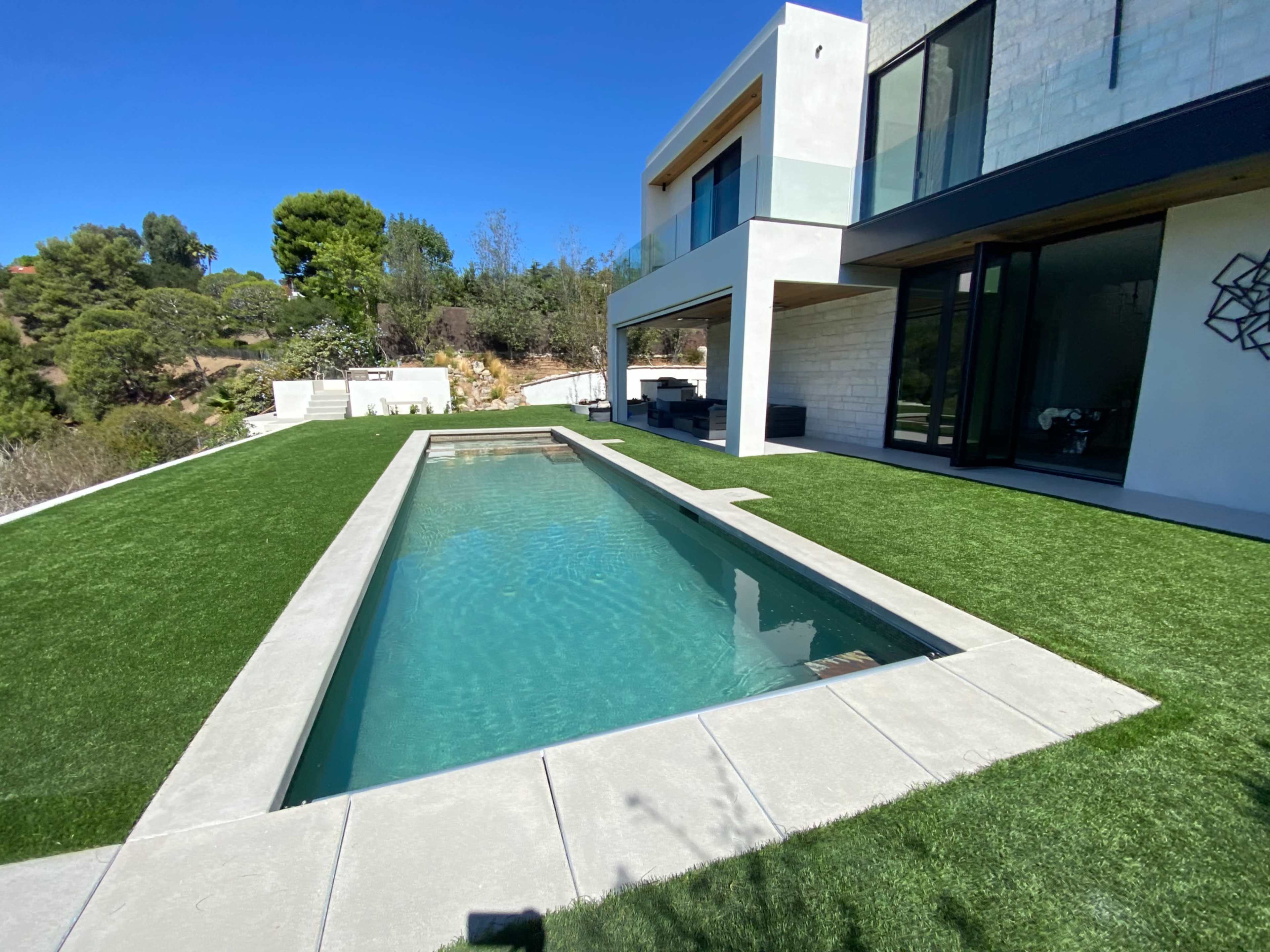 A modern house with large windows beside a rectangular pool surrounded by artificial grass.