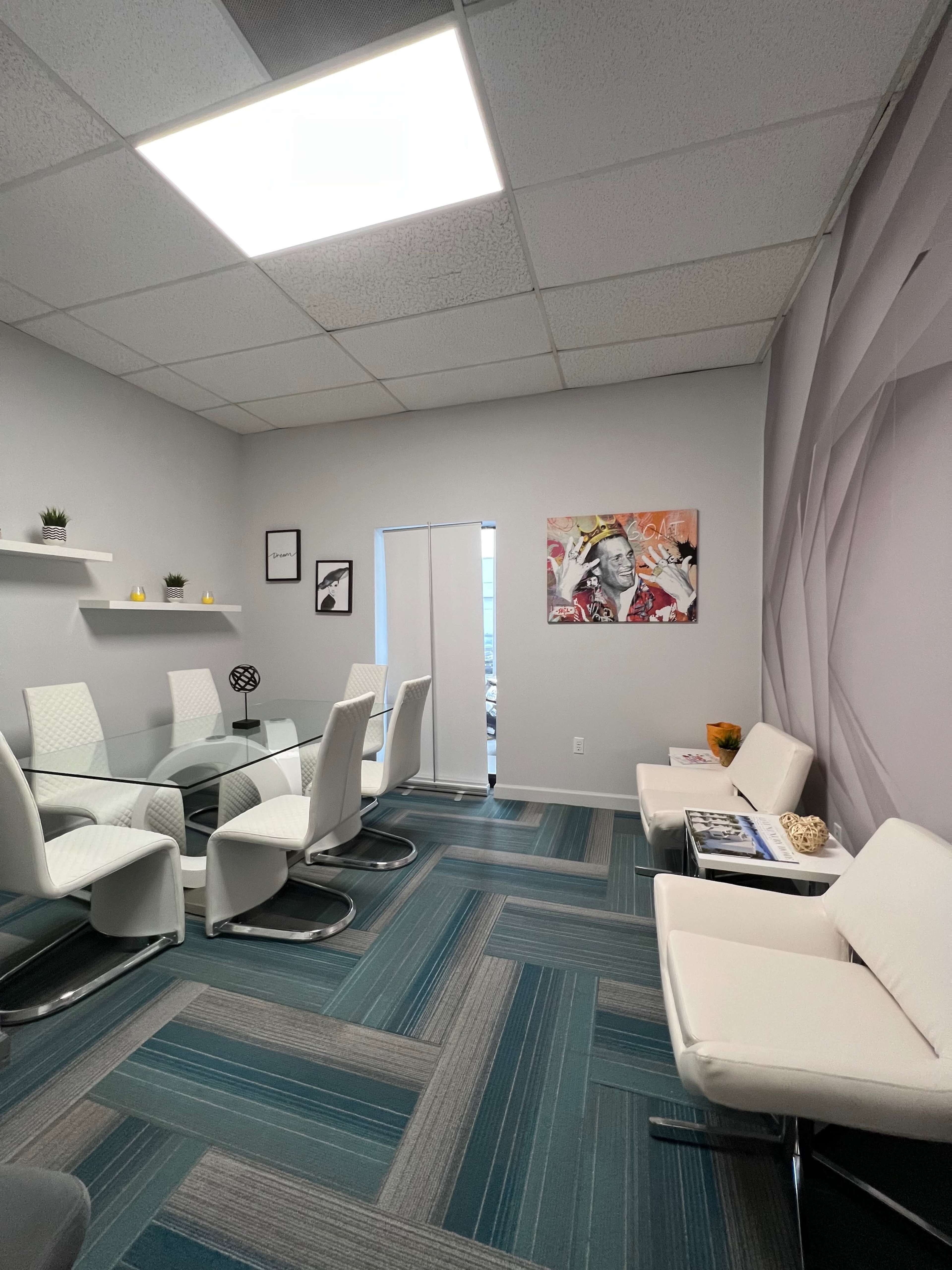 A modern office space with a glass meeting table surrounded by white chairs, light gray walls, and a colorful artwork hanging on one side.