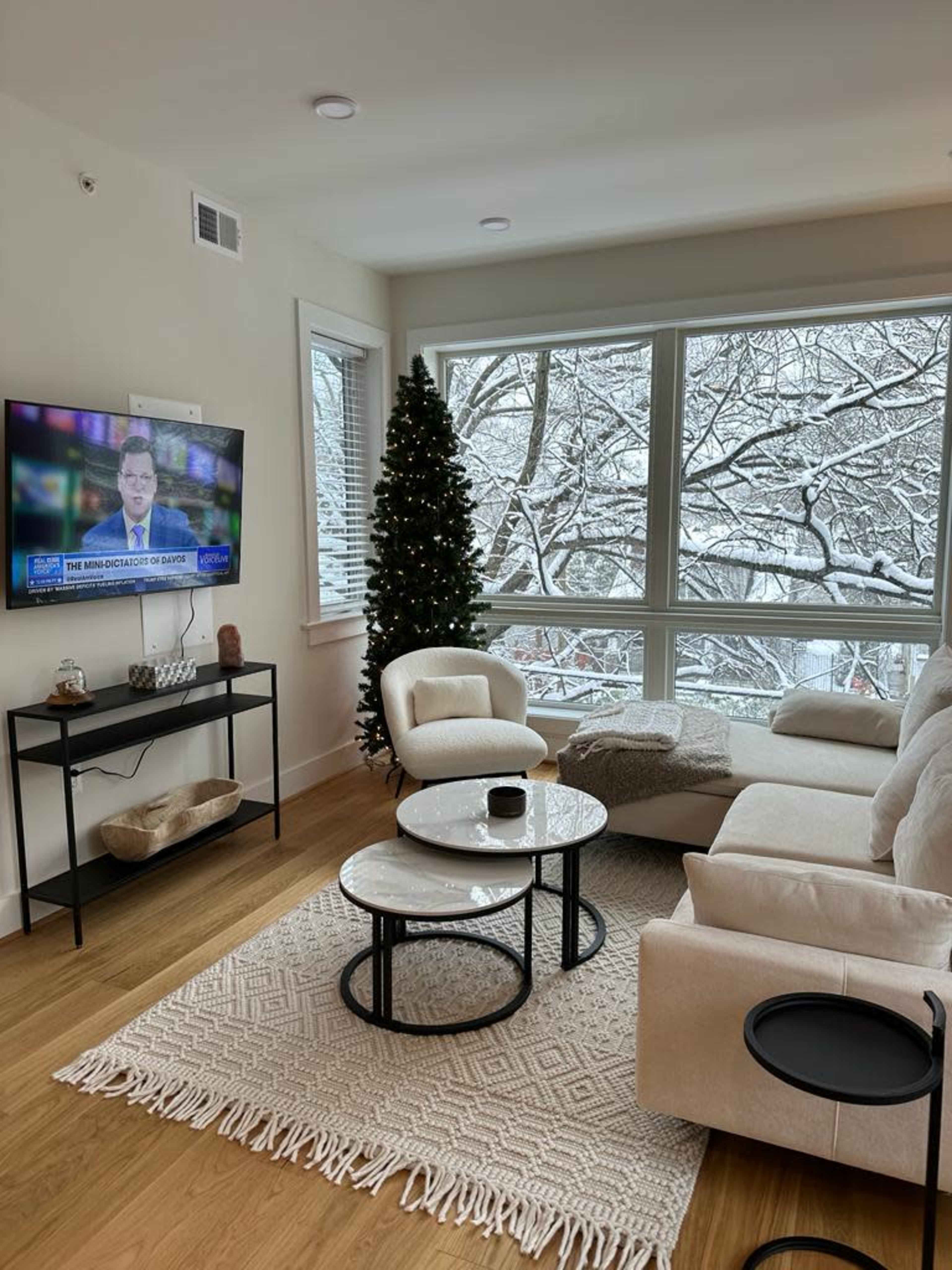 A modern living room with a television displaying news, a Christmas tree, and large windows overlooking a snowy landscape.