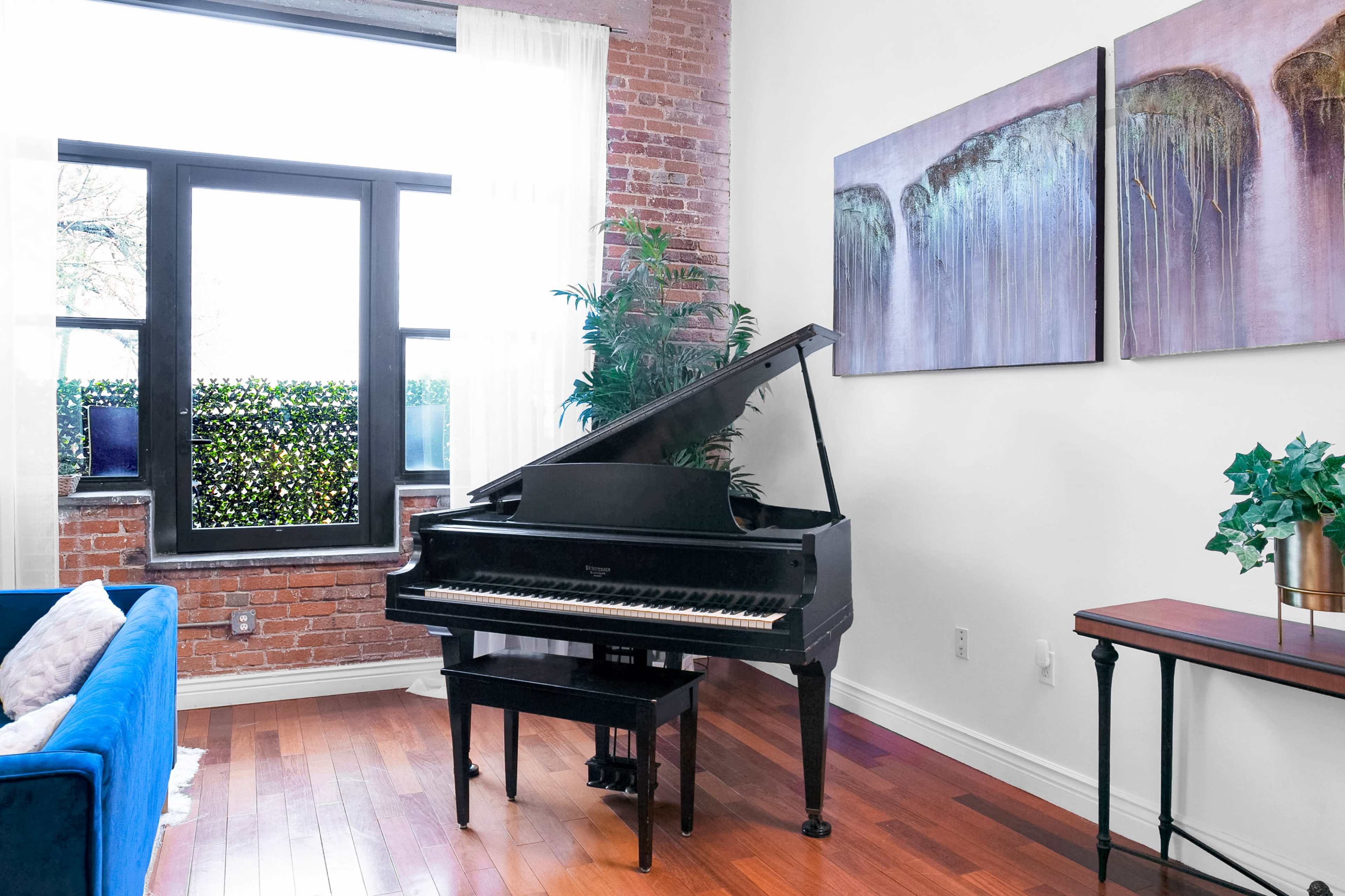 A black grand piano is positioned near a window in a room with brick walls and wooden floors, accompanied by two abstract paintings and a small plant.