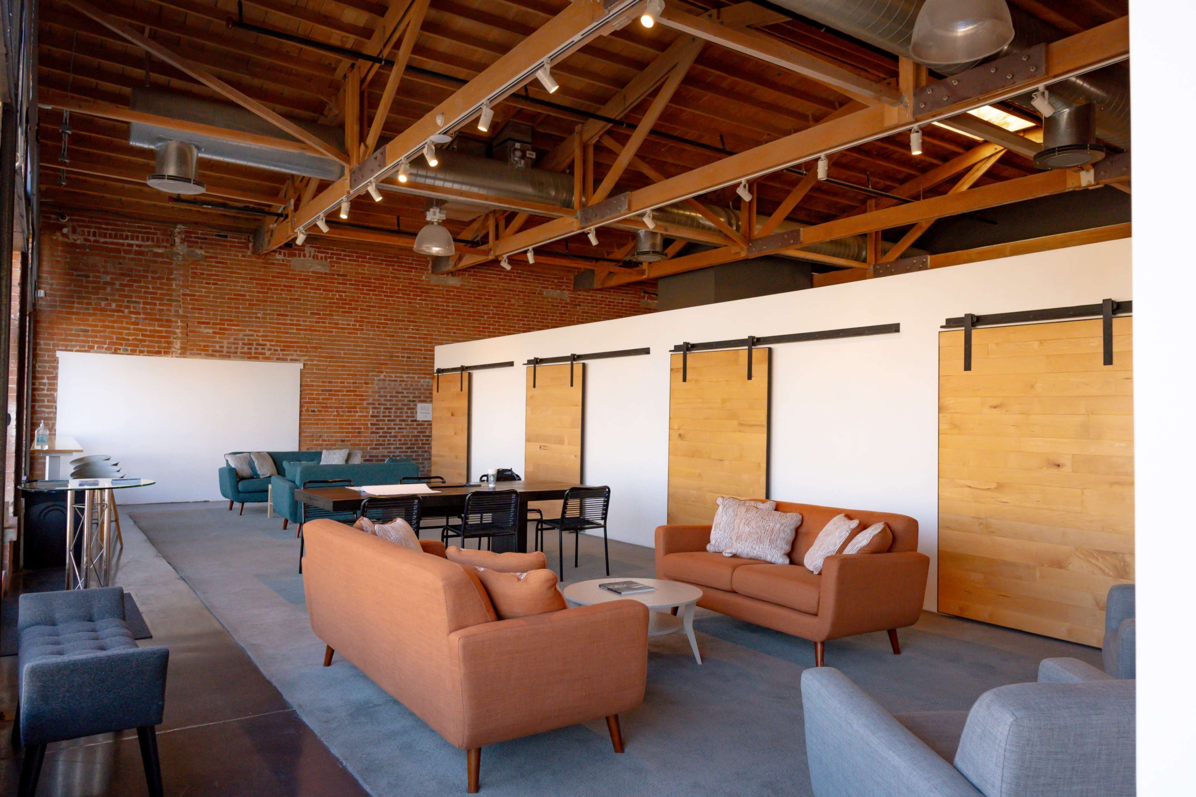 A bright and spacious room featuring wooden beams, a mix of seating options including orange and gray couches, and sliding wooden doors along one wall.