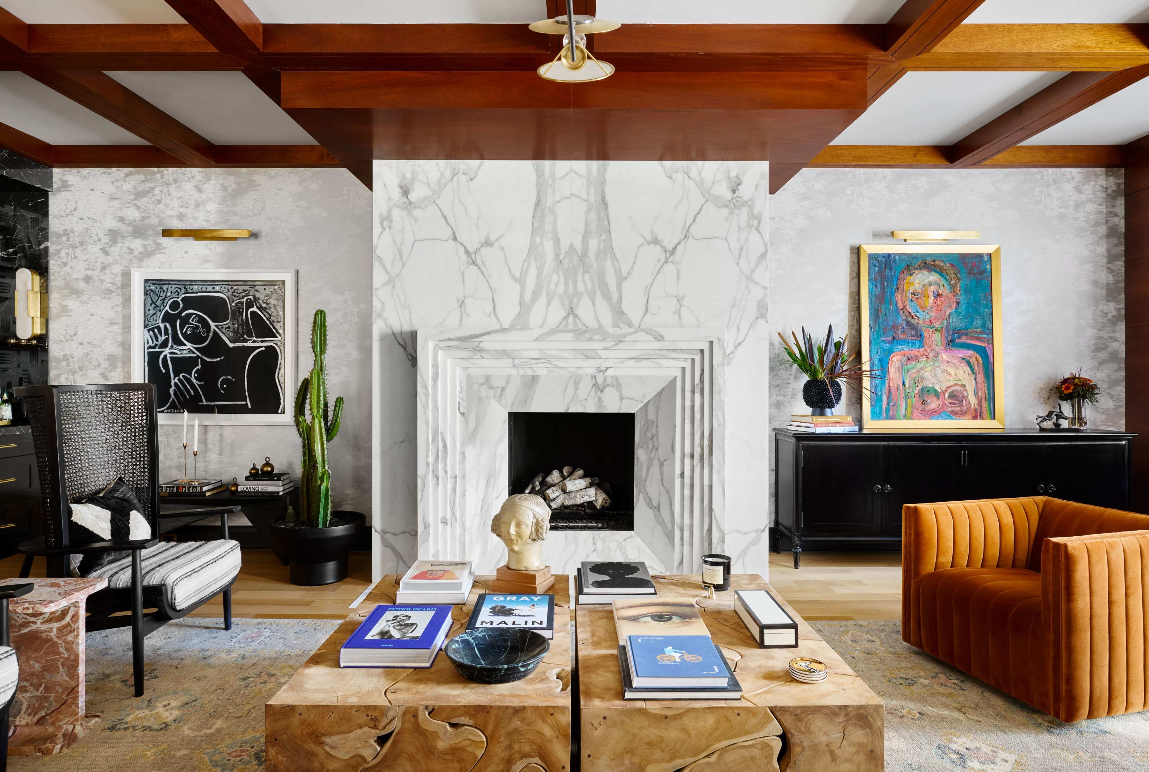A modern living room features a central fireplace with a marble surround, flanked by artwork and plants, with a coffee table adorned with books and decorative items.