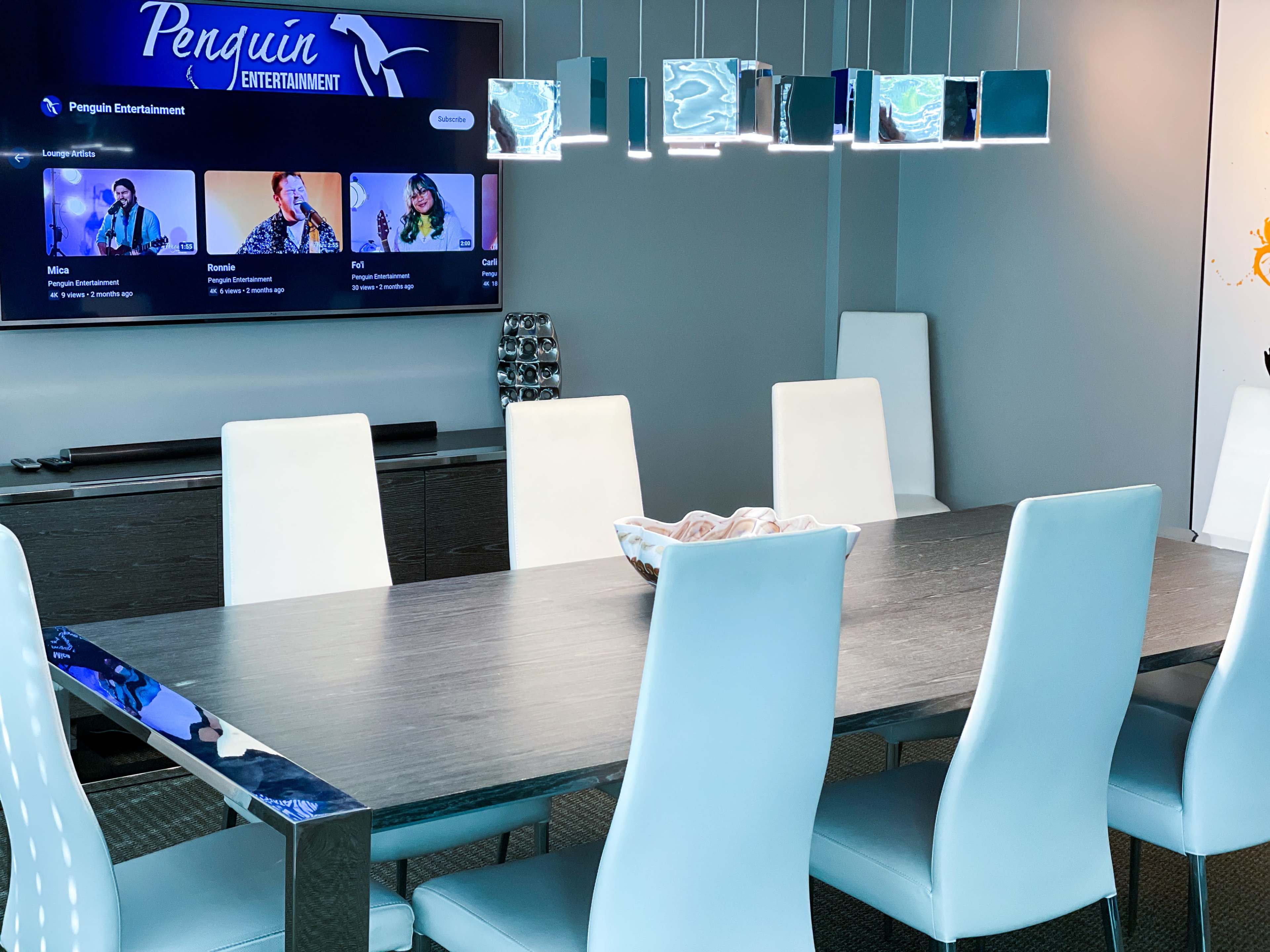 A modern conference room features a long table surrounded by light blue chairs, with a TV displaying "Penguin Entertainment" on the wall.