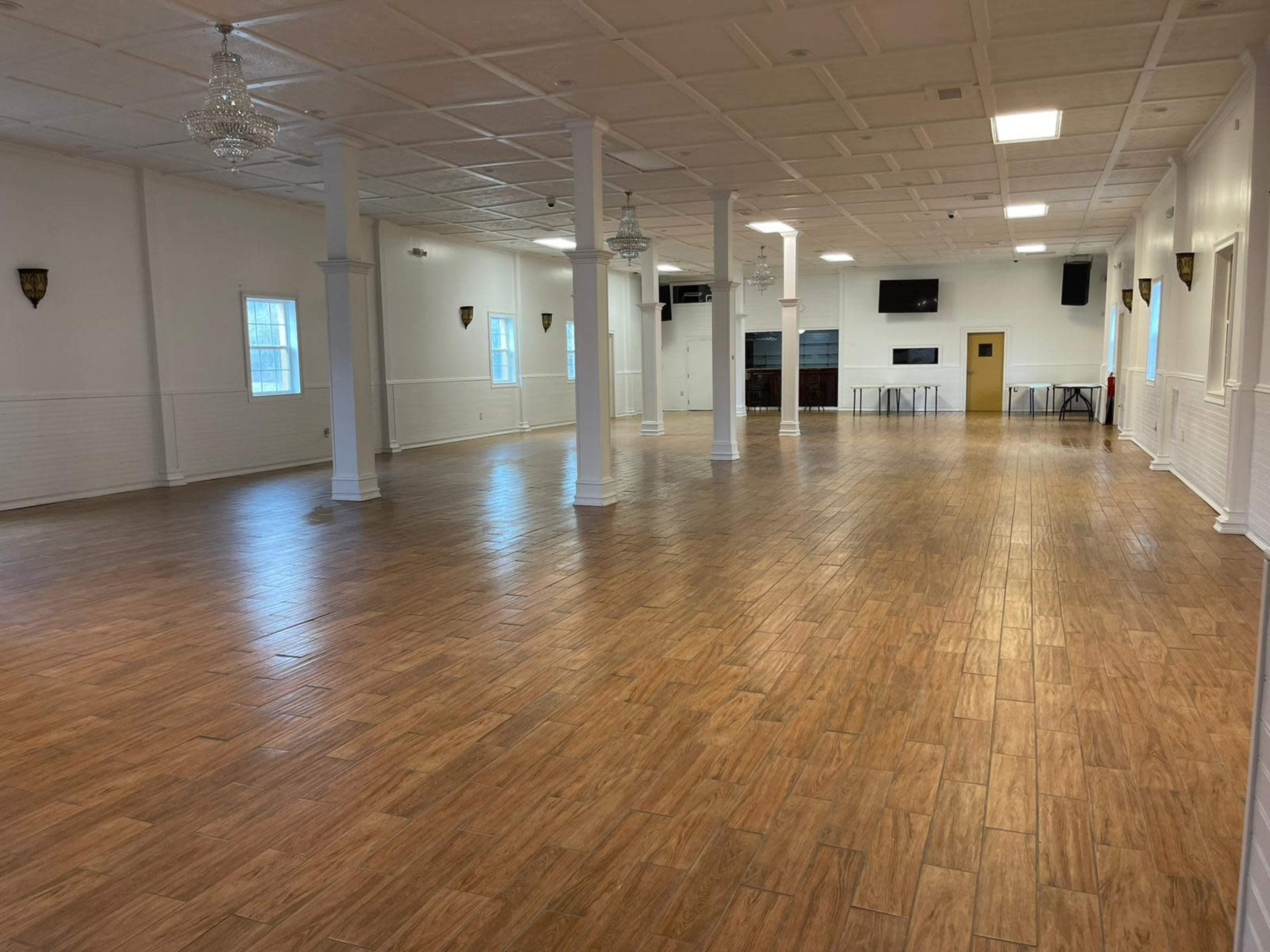The image shows an empty hall with wooden flooring, white walls, and several windows providing natural light.
