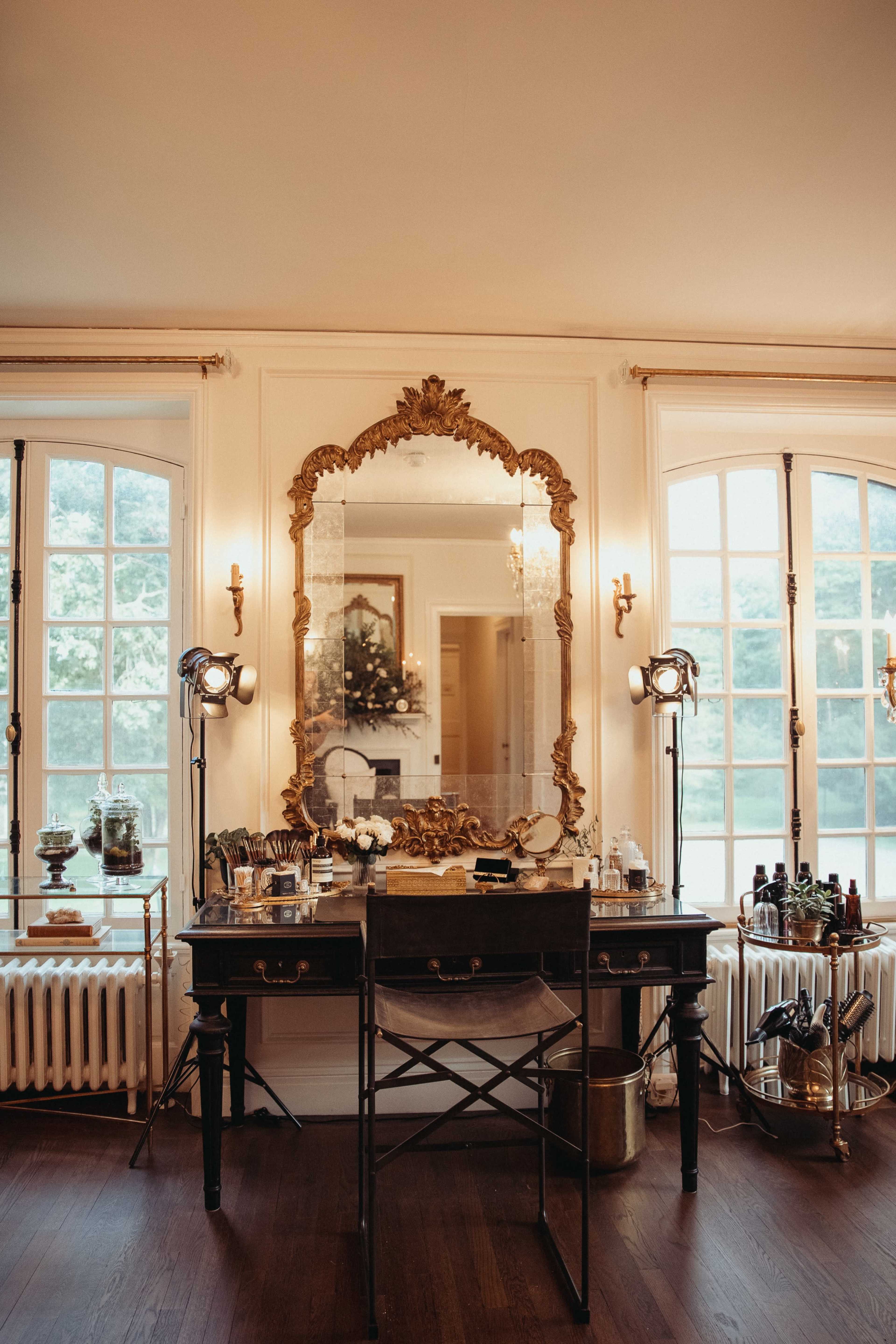 A vintage vanity table with a large ornate mirror is positioned between two windows, surrounded by makeup tools and lighting equipment.