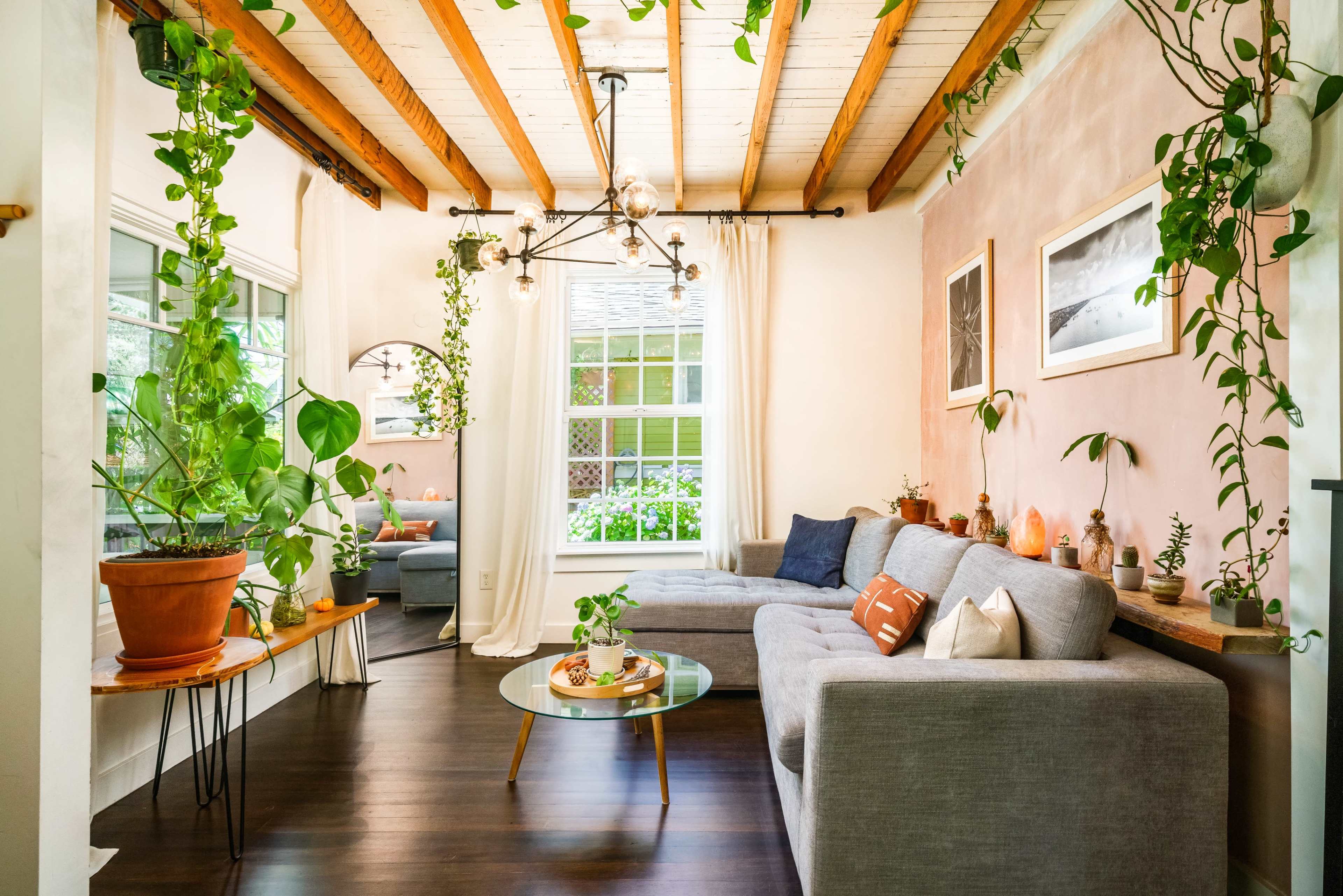 The image shows a well-lit living room featuring a gray sectional sofa, a round coffee table with plants, and large windows with natural light filtering through.
