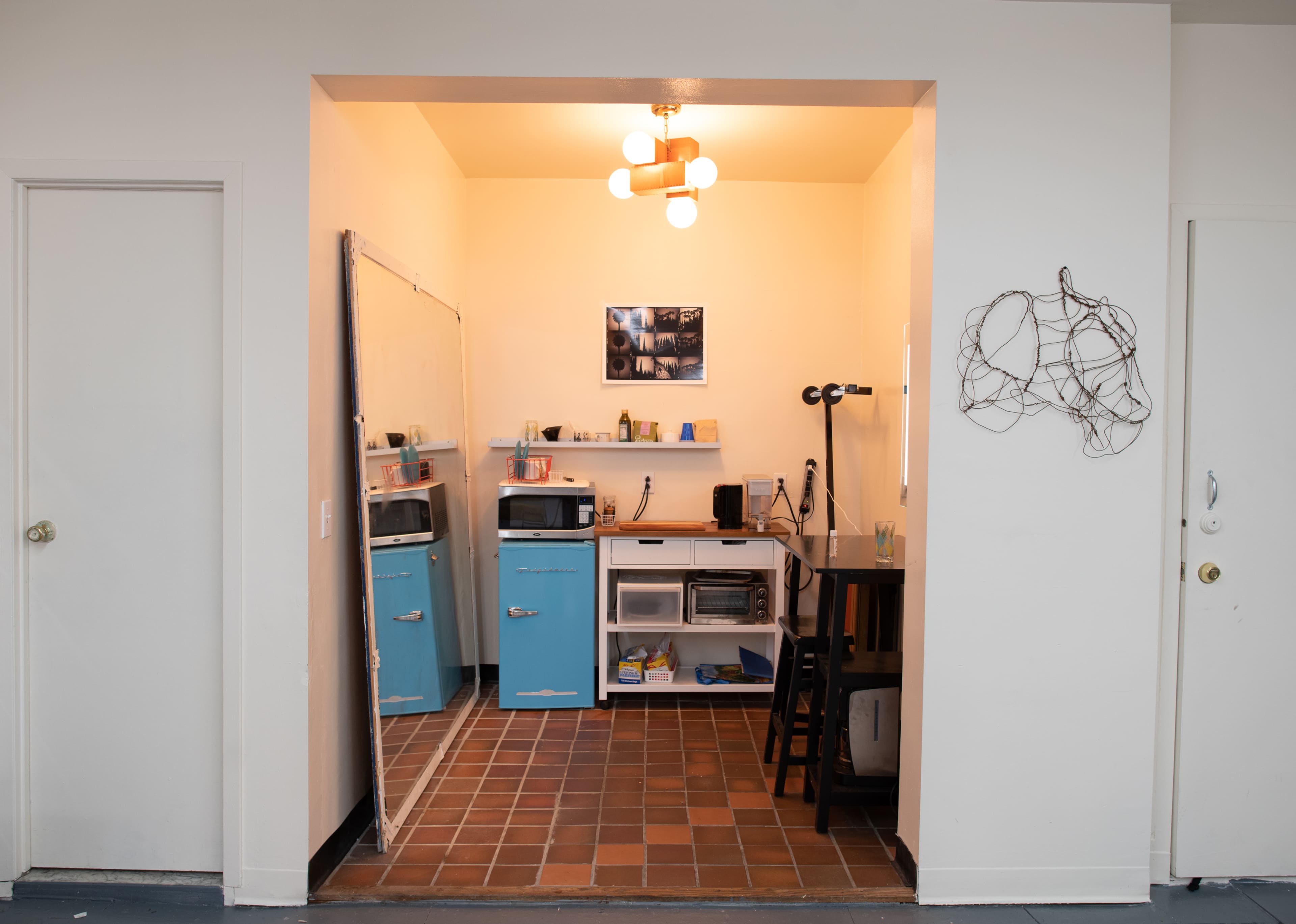 The image shows a small, bright kitchen area featuring light blue cabinets, a mirror, and a tiled floor, with a table and chair set against the wall.