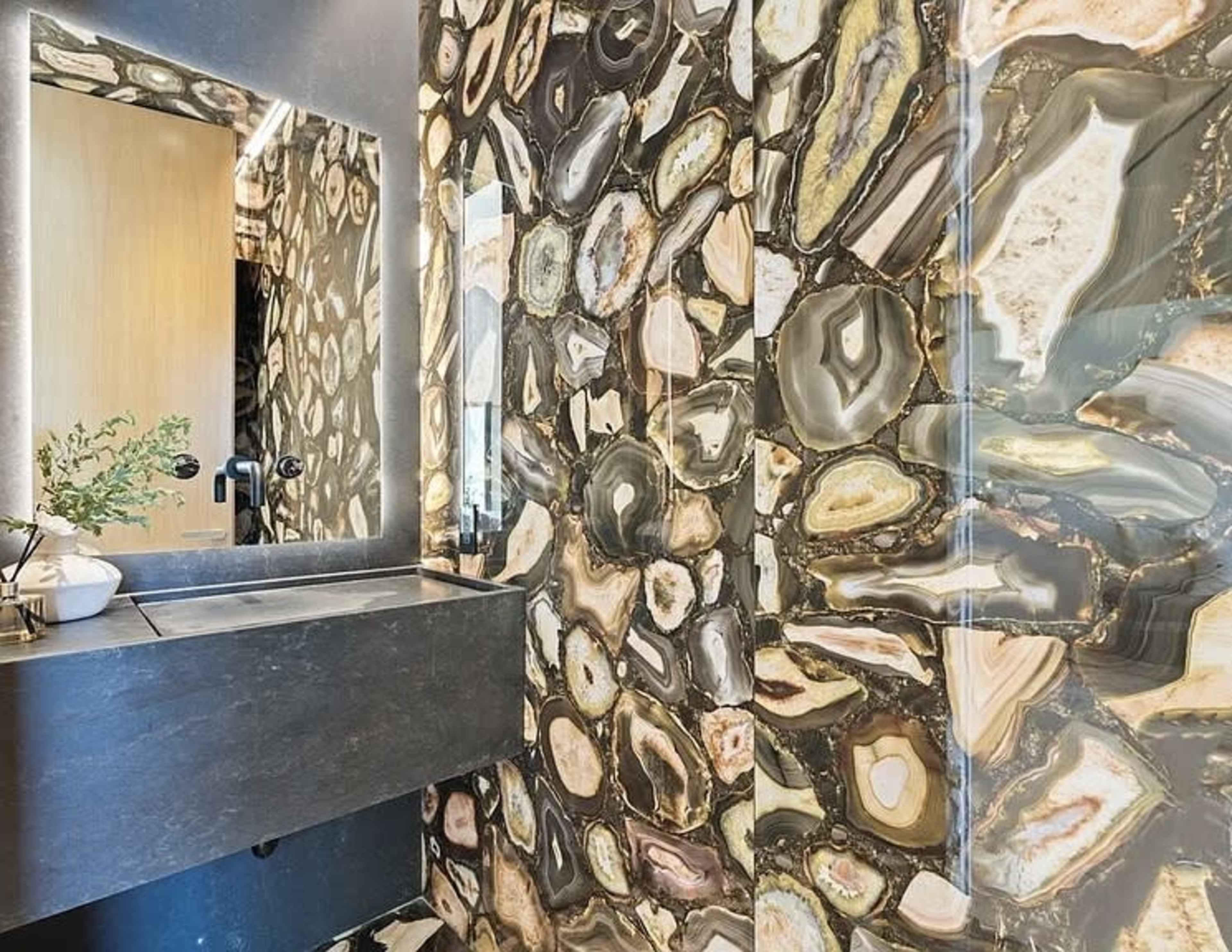 The image features a bathroom with a striking wall design made of colorful, patterned stone slabs and a sleek black countertop with a mirror above it.